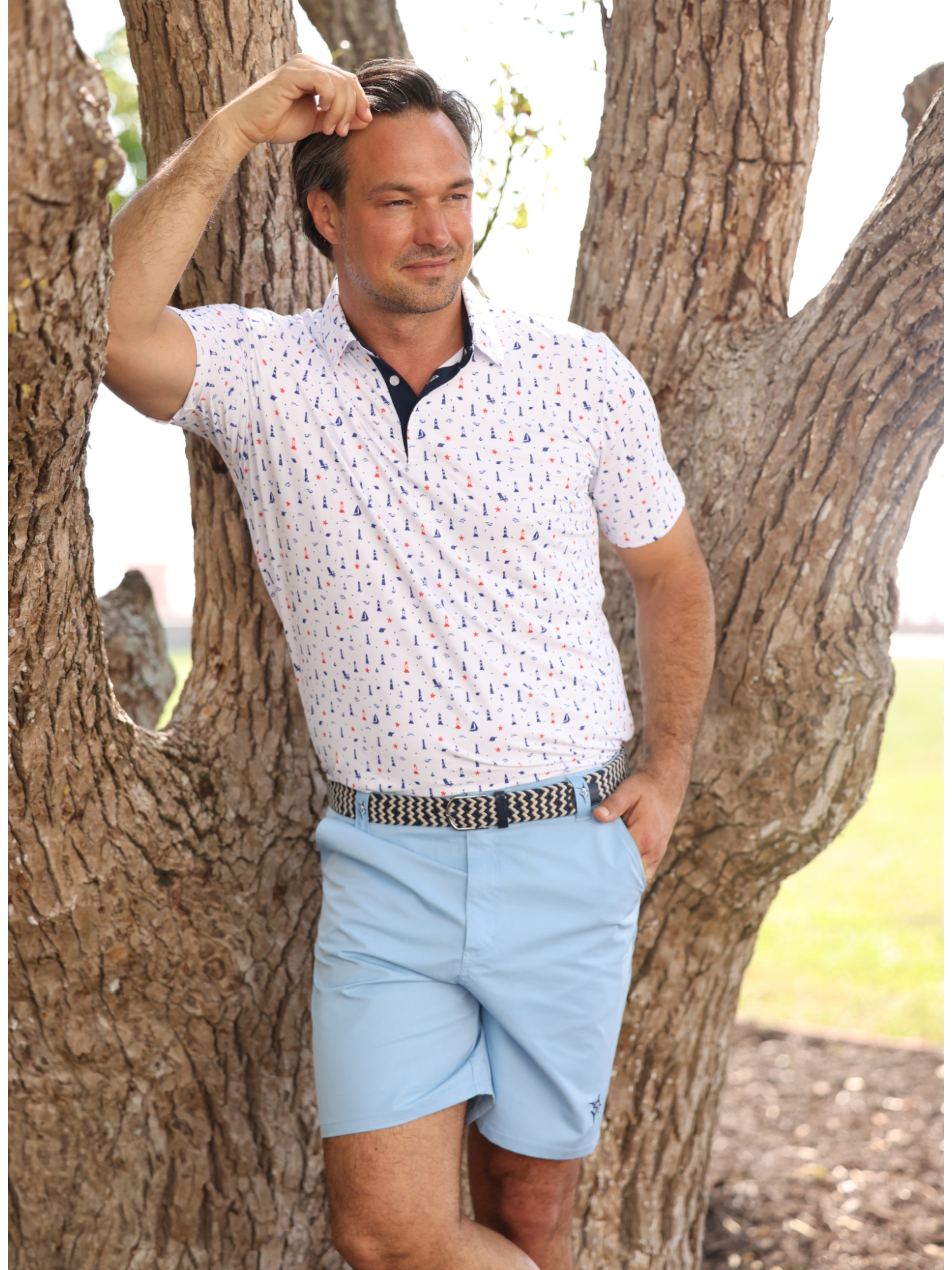 Man wearing a patterned shirt and light blue shorts standing against a tree