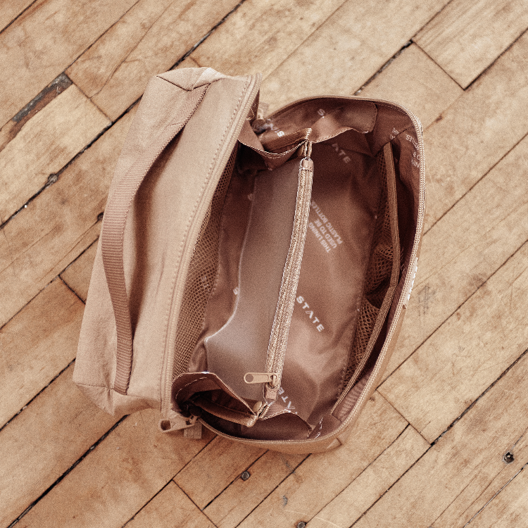 A beige canvas zippered pouch with a matching fabric handle, shown open from a top-down view on a rustic wooden plank floor. The interior features a metallic bronze-brown lining with a zippered compartment and mesh pockets.