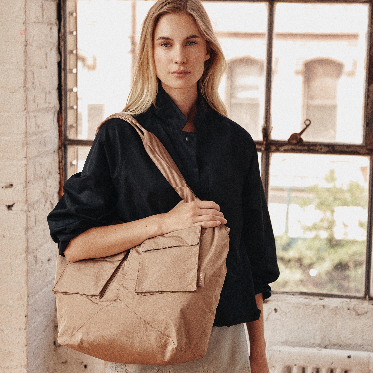 A woman wearing a black shirt and light-colored pants carries a large tan canvas tote bag with multiple front pockets and a wide shoulder strap, standing in front of a bright window with a brick wall on the left.