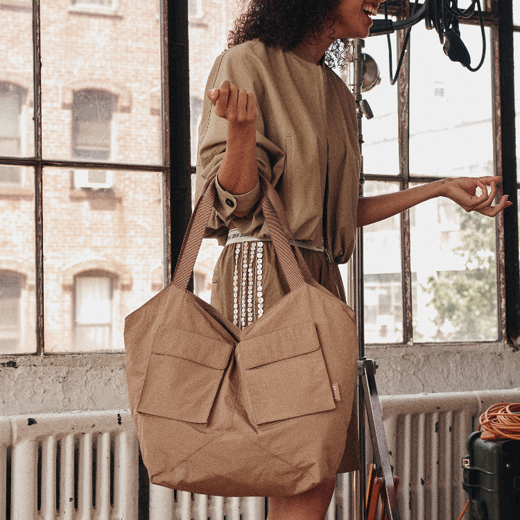 A woman in a beige dress carries a large tan canvas tote bag with two front flap pockets, standing in front of a window with a brick building outside.