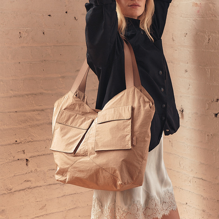 A woman wearing a black button-up shirt and white lace-trim shorts carries a large tan canvas shoulder bag with two front flap pockets against a textured beige brick wall.