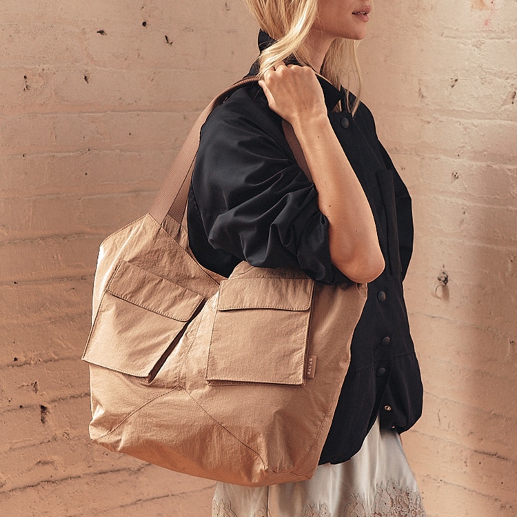 A woman wearing a black button-up shirt and white skirt carries a large tan canvas tote bag with two front flap pockets, posed against a textured beige brick wall.