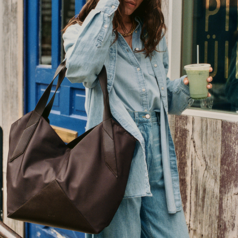 Person holding a brown bag and a drink, wearing a denim outfit, standing in front of a blue door.