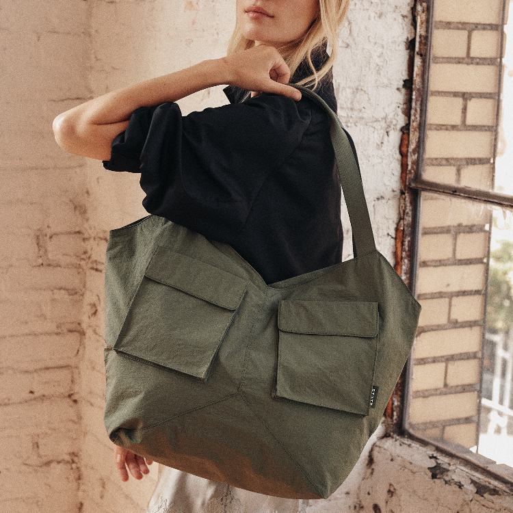 A woman wearing a black shirt carries a large olive green canvas tote bag with two front flap pockets, standing beside a brick wall and window.