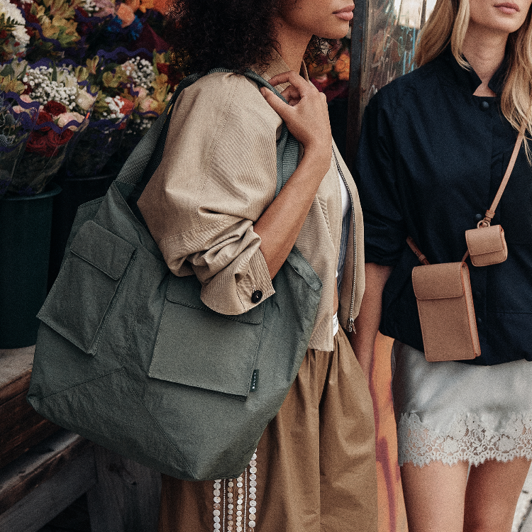 A woman wearing a beige shirt and brown skirt carries a large olive green canvas tote bag over her shoulder in a rustic, flower-filled setting. Another woman in a black shirt and white skirt is partially visible on the right carrying a small tan crossbody pouch.