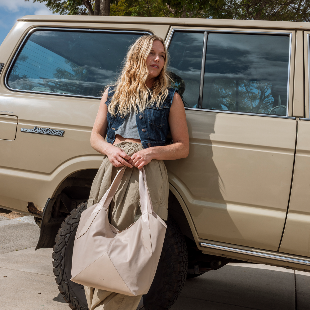 Woman leaning against a beige Land Cruiser with a large beige tote bag.