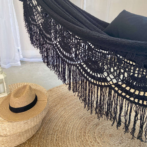Black macramé hammock with fringes on a textured surface, next to a straw hat.