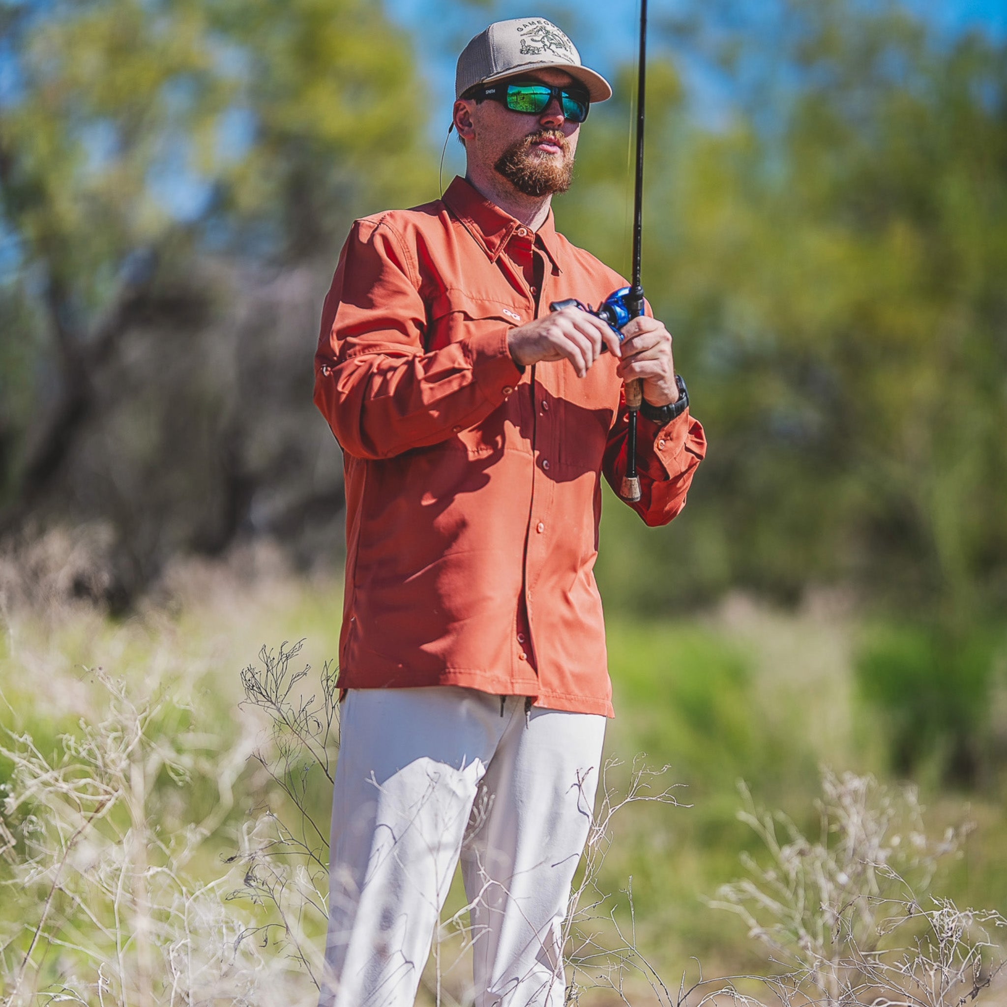Man in an orange jacket and sunglasses holding a fishing rod in a natural setting