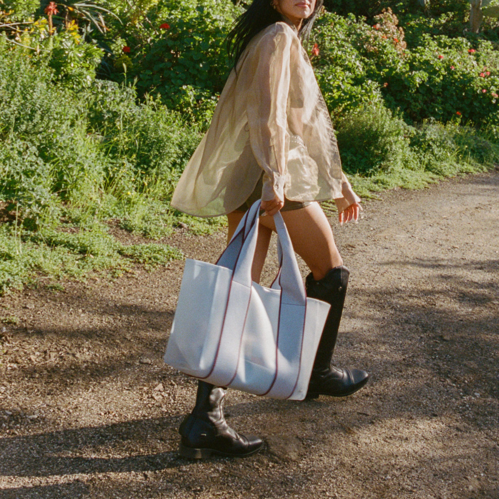 Woman walking outdoors holding a white tote bag with red stripes