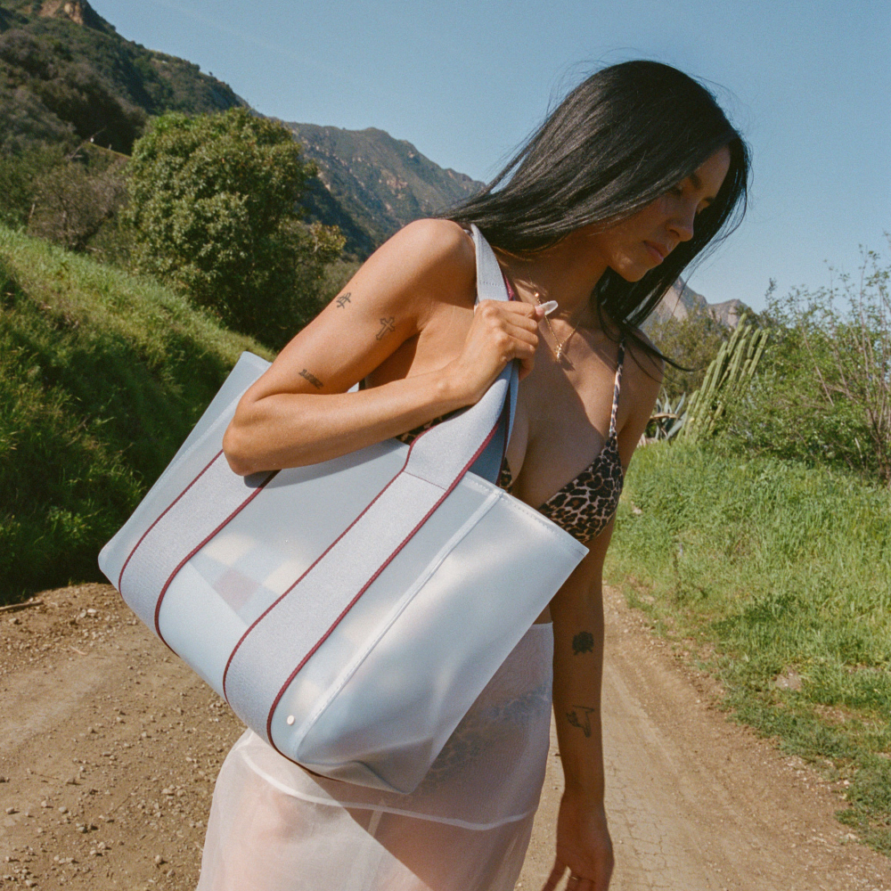 Woman holding a large white tote bag with red stripes in a natural setting with mountains and greenery.