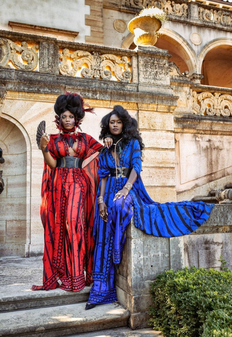 Two women in vibrant red and blue dresses standing on stone steps with classical architecture in the background.