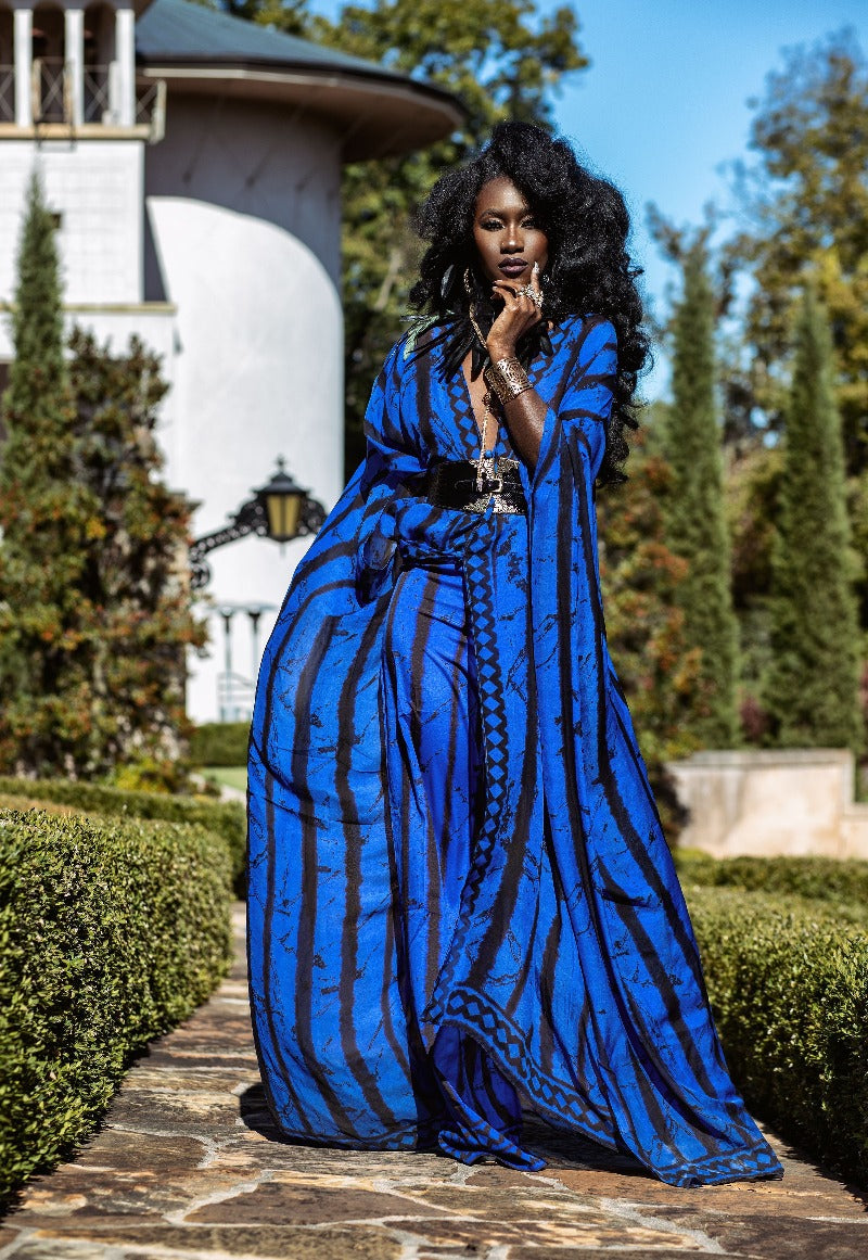 Woman in a blue dress standing outdoors with greenery and a building in the background