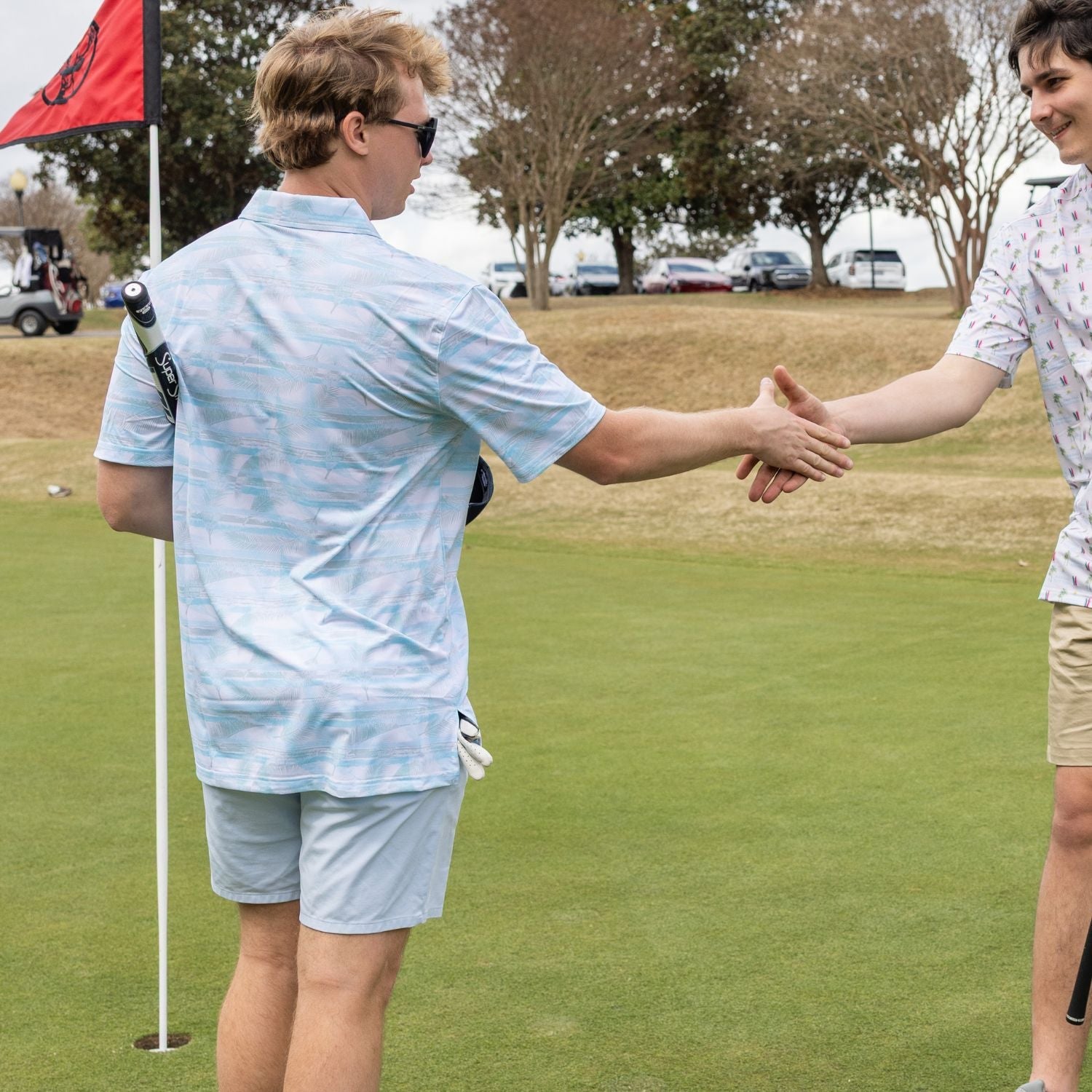 Two people shaking hands on a golf course with trees and cars in the background.