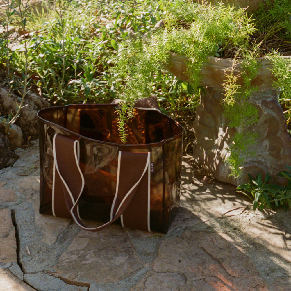 Brown leather tote bag with white straps on a stone surface with greenery