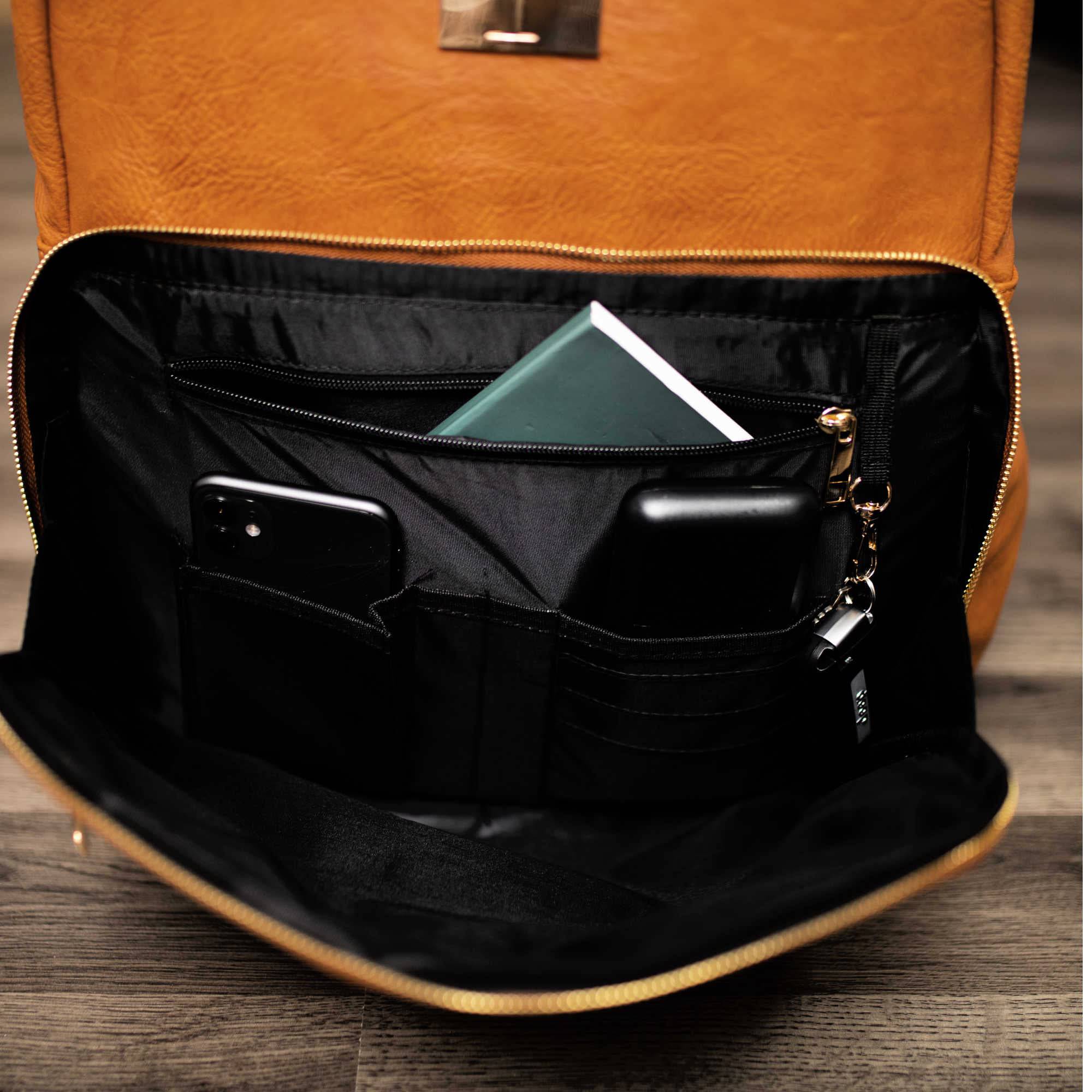 Brown leather backpack with open front pocket showing phone, wallet, and notebook on a wooden surface.