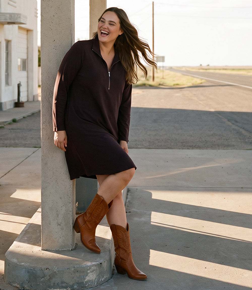 A woman wearing a dark brown long-sleeve midi dress with a half-zip collar and brown cowboy boots, leaning against a concrete pole on a sunny road.