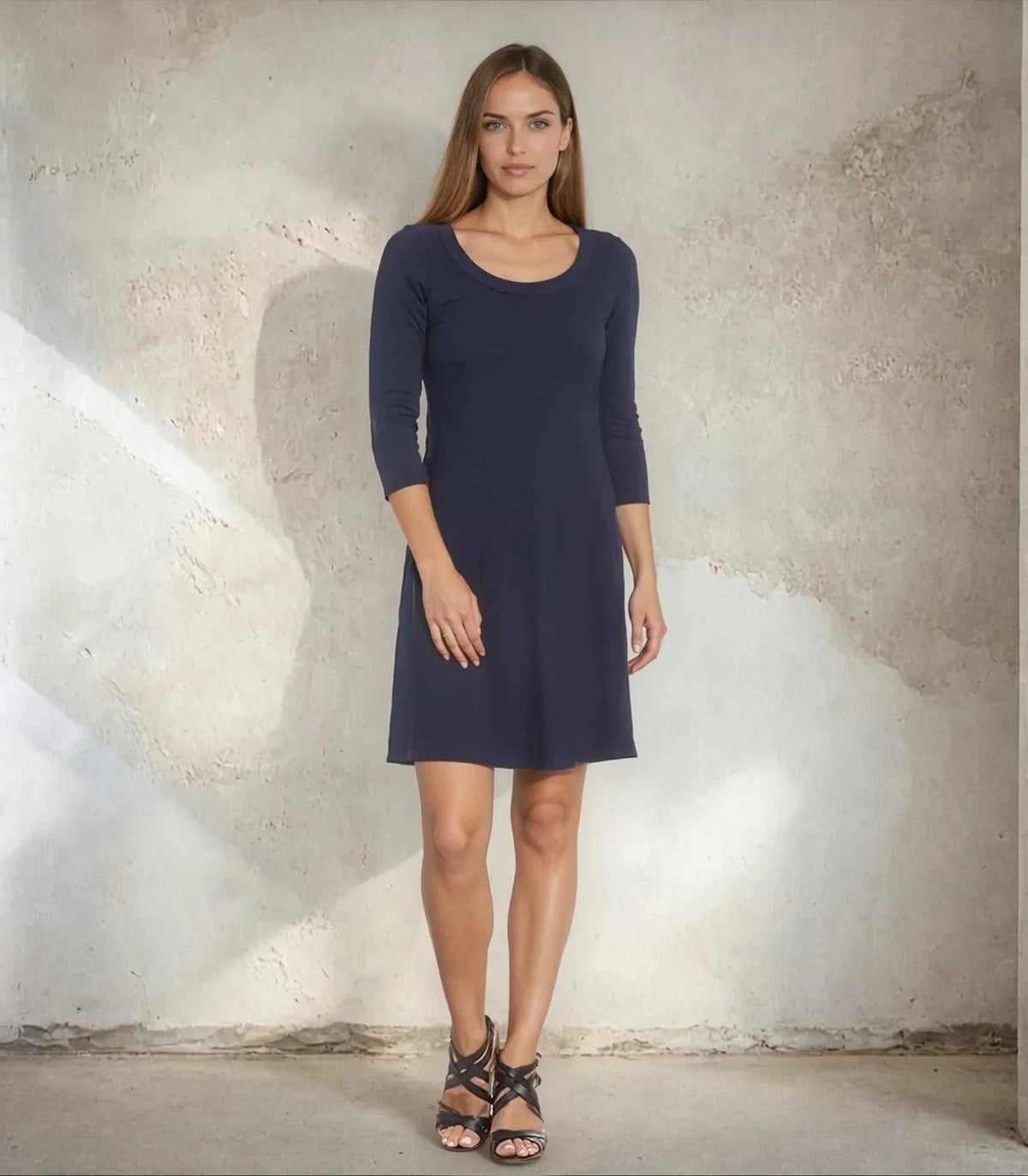 A woman modeling a navy blue three-quarter sleeve shift dress with a scoop neckline, standing against a textured concrete wall.