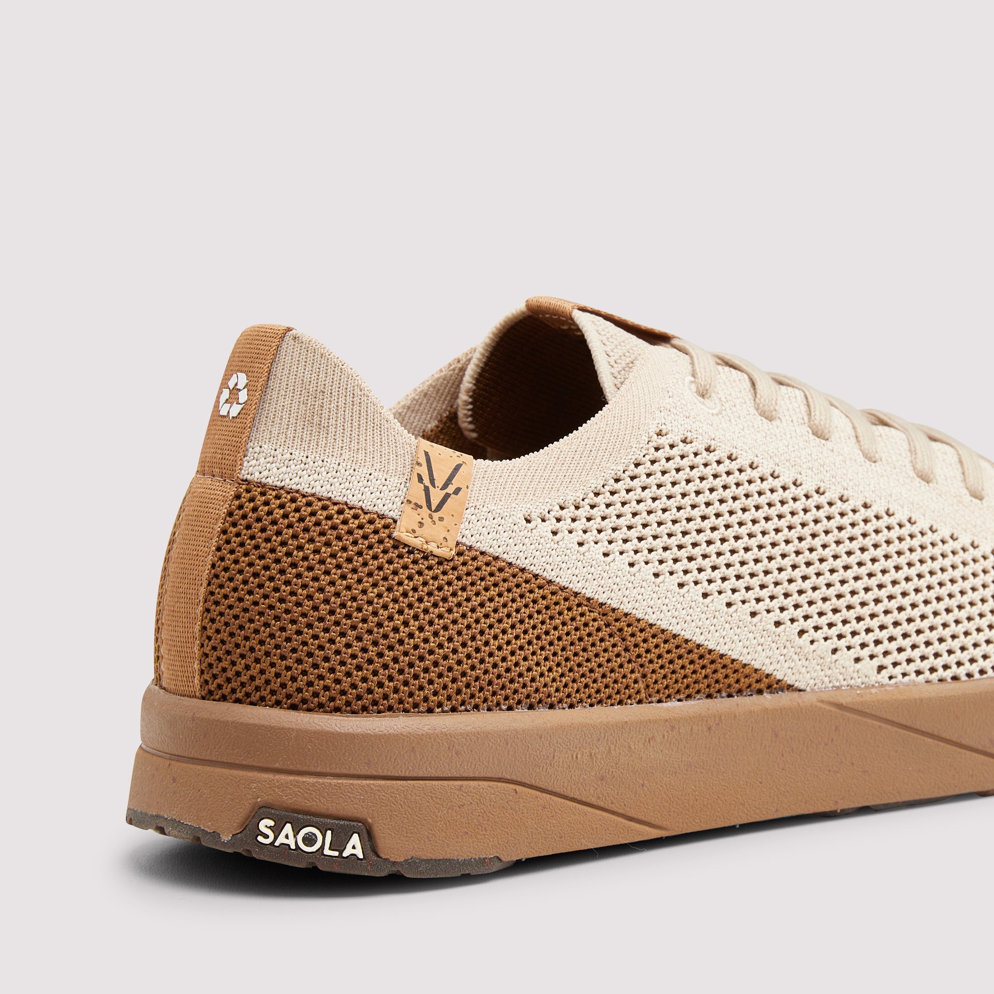 A close-up side view of a knit sneaker in beige and brown, featuring a textured knit upper, perforated details, a brown midsole, and a small SAOLA logo on the heel.