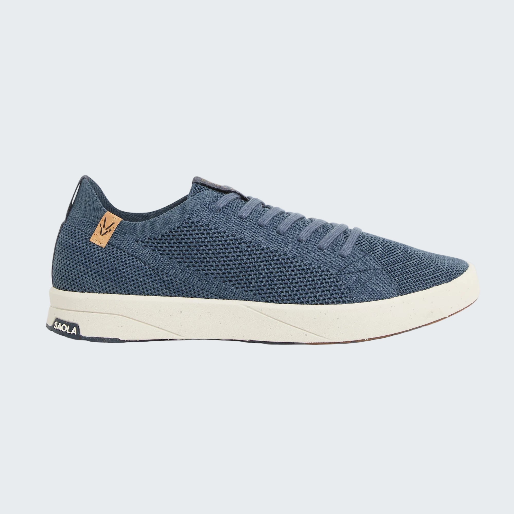 A single low-top sneaker shown in profile against a light gray background. The shoe features a textured navy blue mesh upper, matching navy laces, a cream-colored midsole, and a small tan leather heel tab with a stylized 'V' logo.