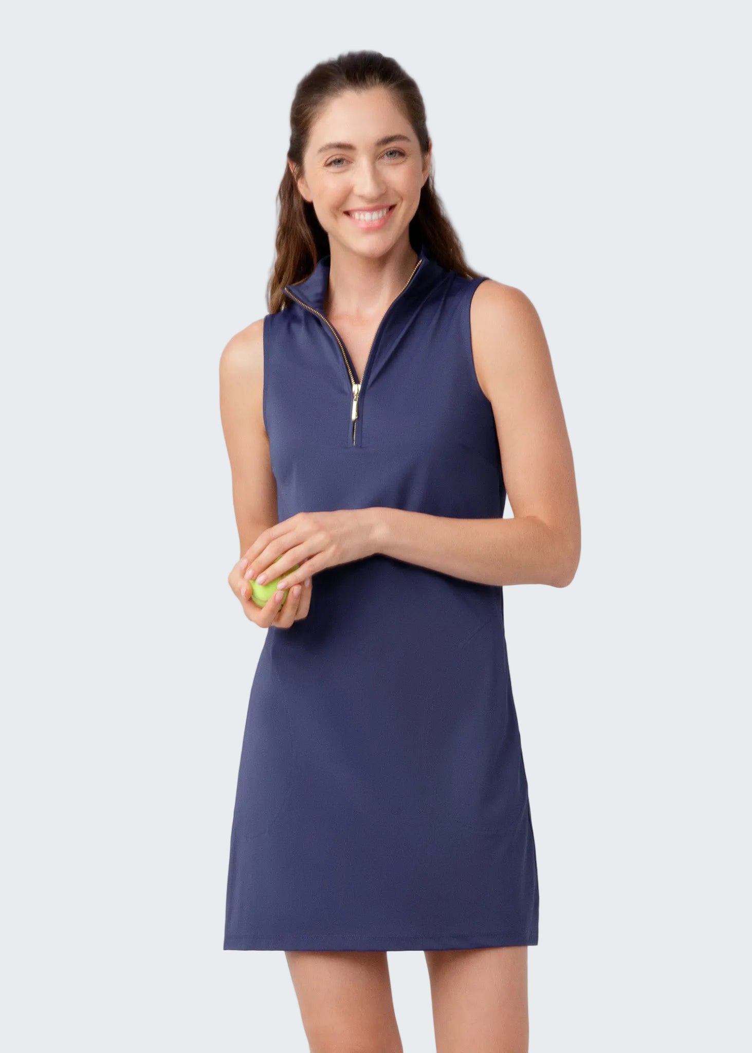 Woman wearing a navy sleeveless dress with a zipper, holding a tennis ball on a light gray background
