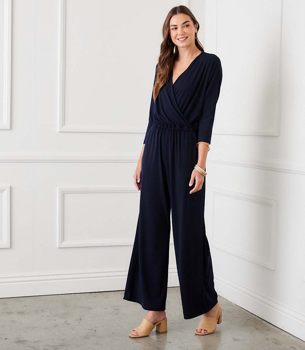 A woman modeling a navy blue wrap-style wide-leg jumpsuit with three-quarter sleeves, standing in a bright room with white paneled walls and a gray floor.