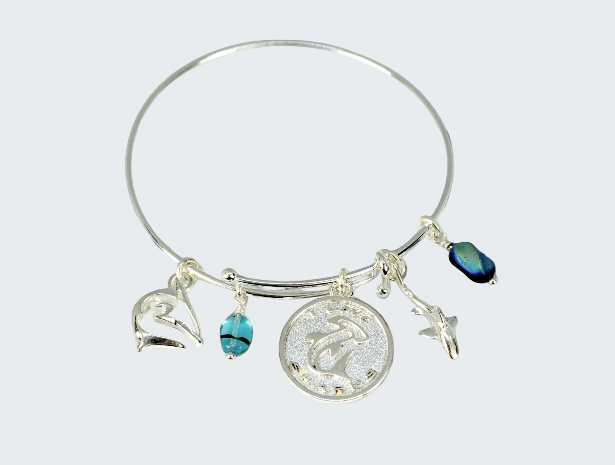 A silver bangle bracelet with sea-themed charms, including a dolphin pendant, a seashell charm, a starfish charm, and blue beads, displayed on a weathered wooden surface.