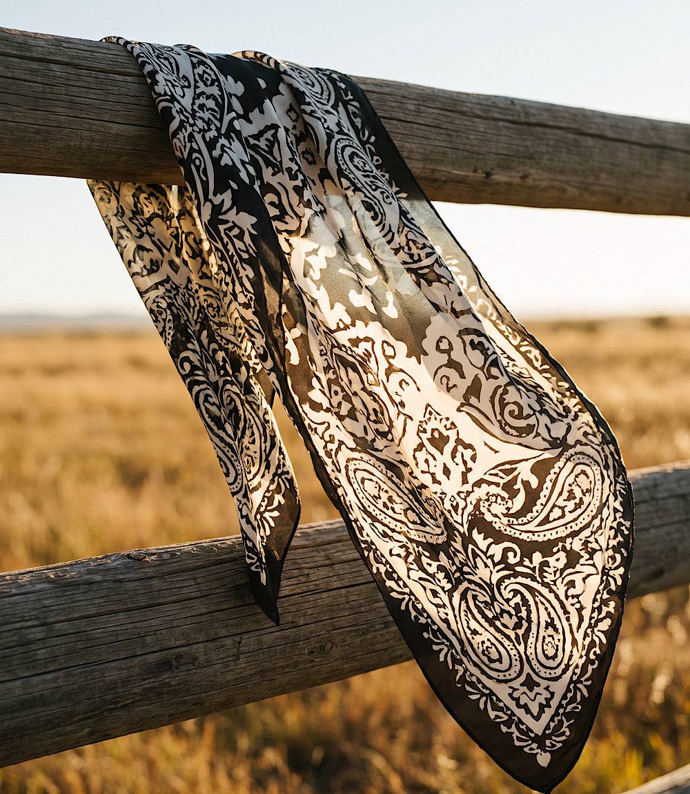 A sheer black and beige paisley bandana scarf draped over rustic wooden fence rails in an outdoor field at sunset.