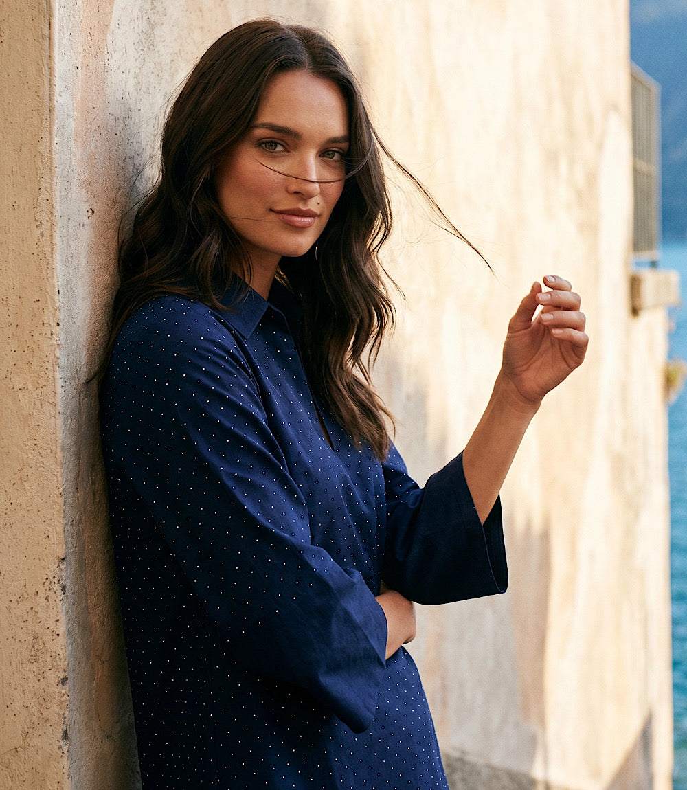 Woman in a blue dress leaning against a textured wall with sunlight filtering through.