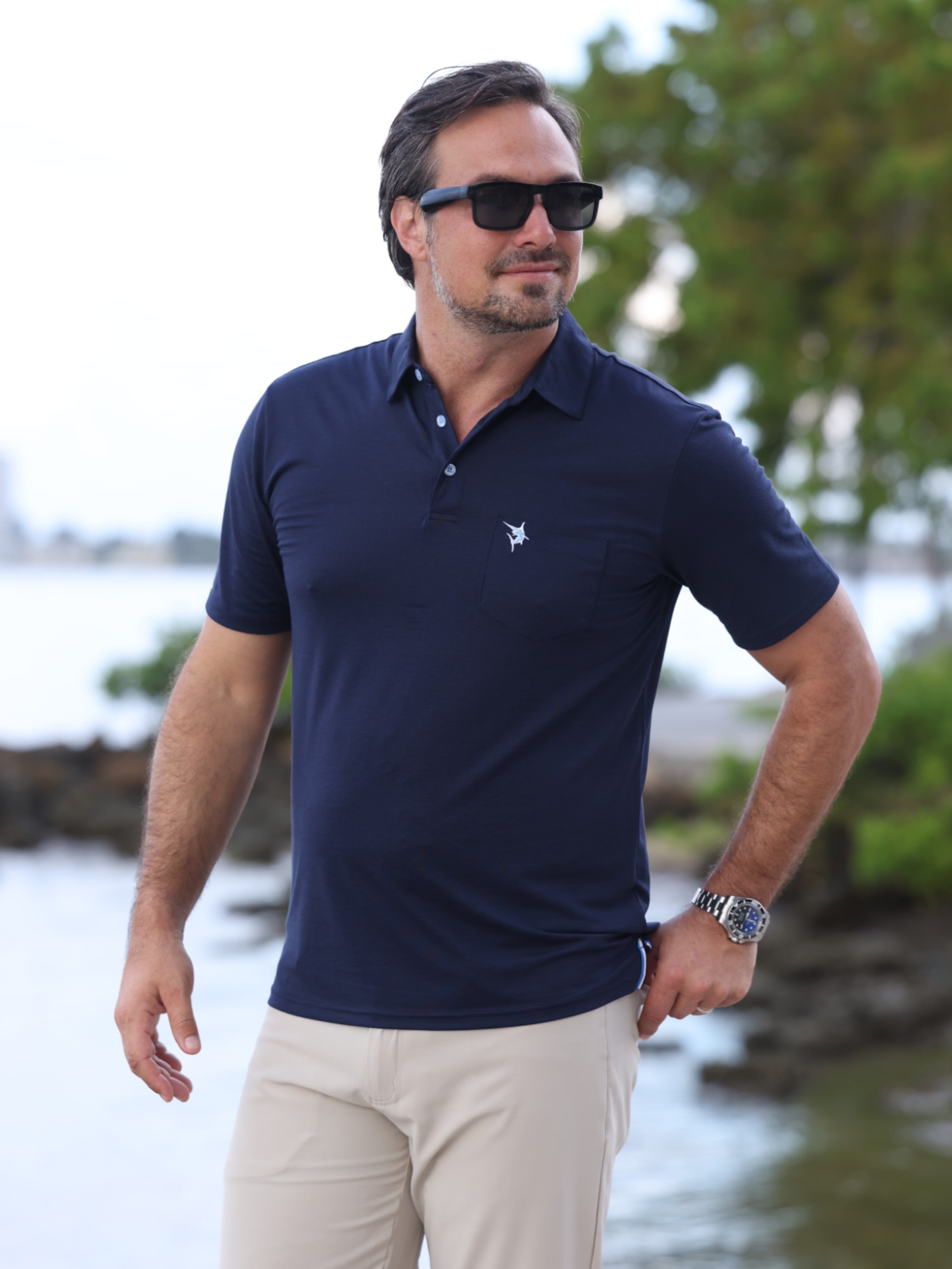 Man wearing a navy polo shirt with a logo, sunglasses, and beige pants by a water body.
