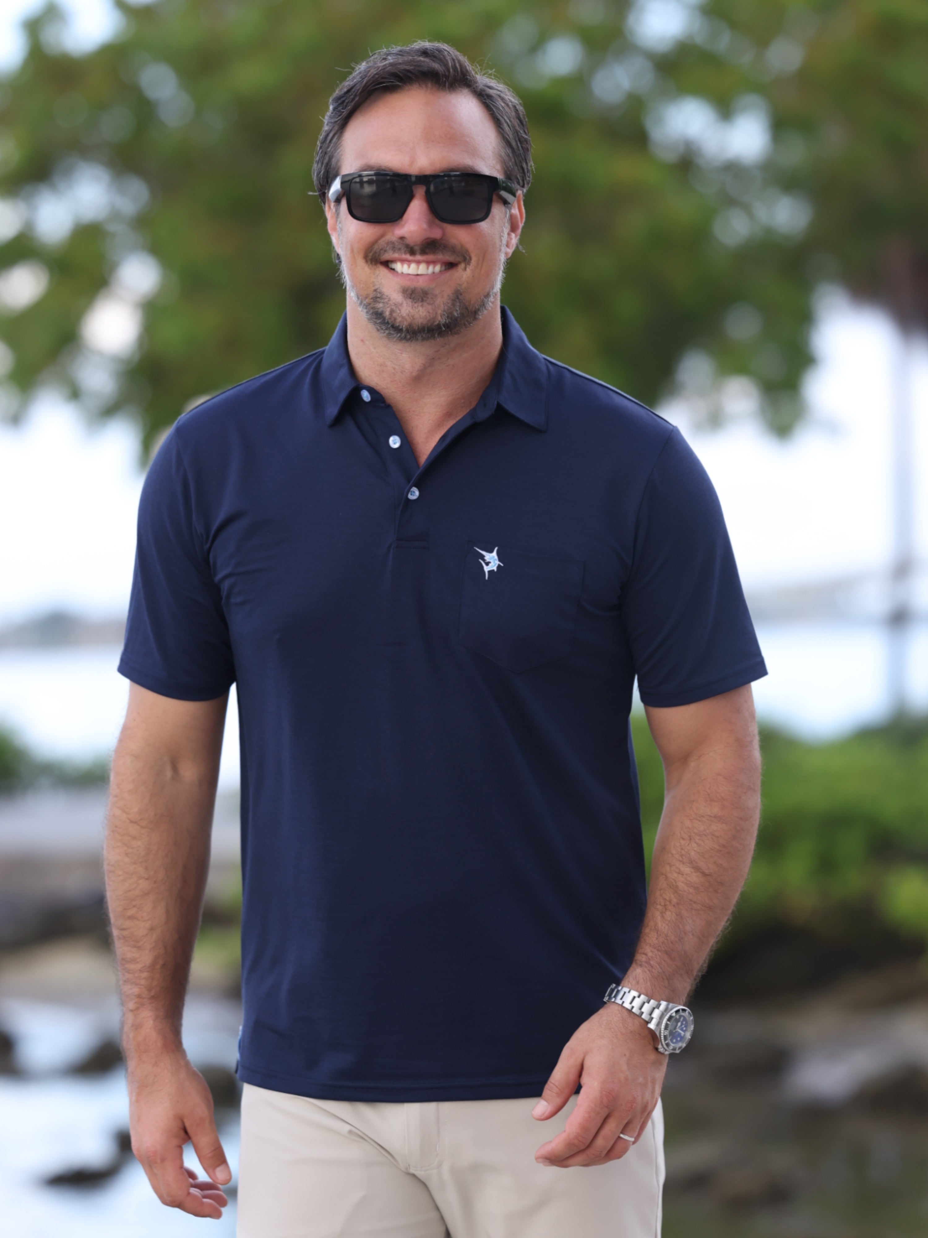 Man wearing a navy polo shirt with a logo, sunglasses, and beige pants outdoors.