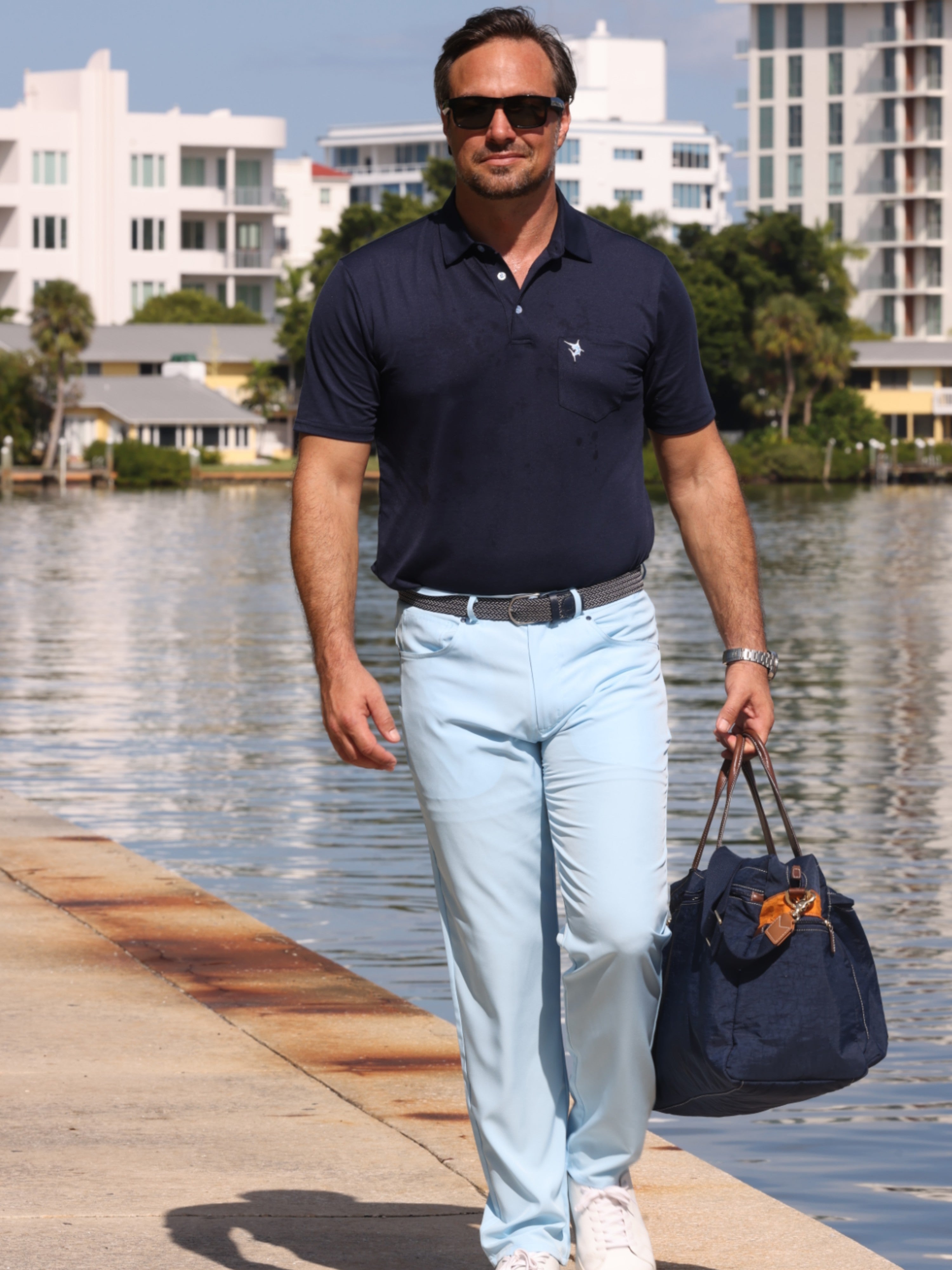 Man in navy polo shirt and light blue jeans holding a navy bag by a waterfront.