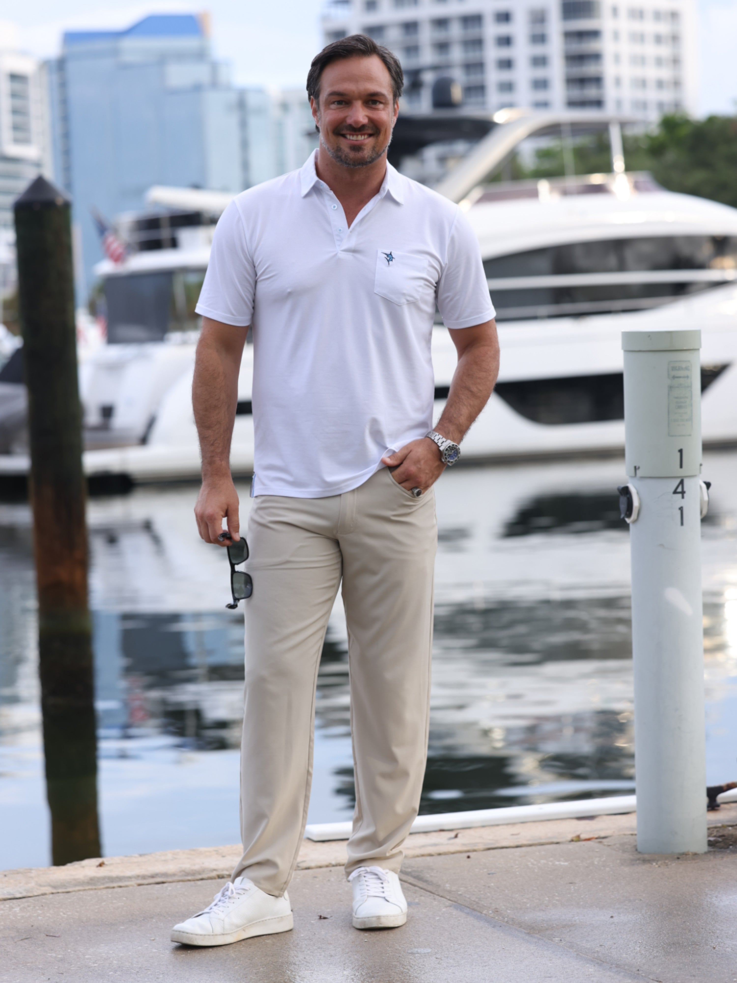 Man in a white polo shirt and beige pants standing by a marina with boats in the background.