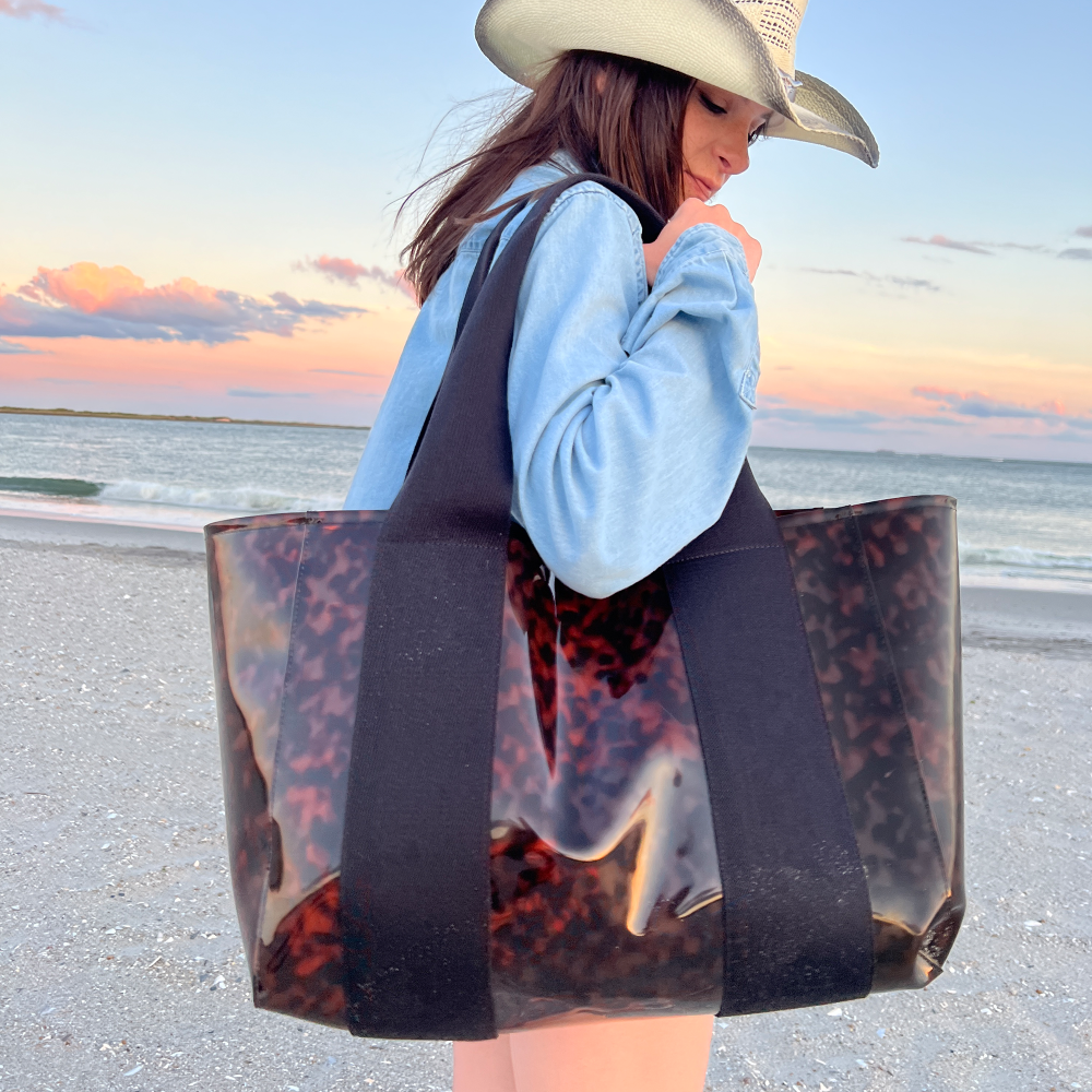 Person holding a transparent tote bag with tortoiseshell pattern on a beach at sunset.