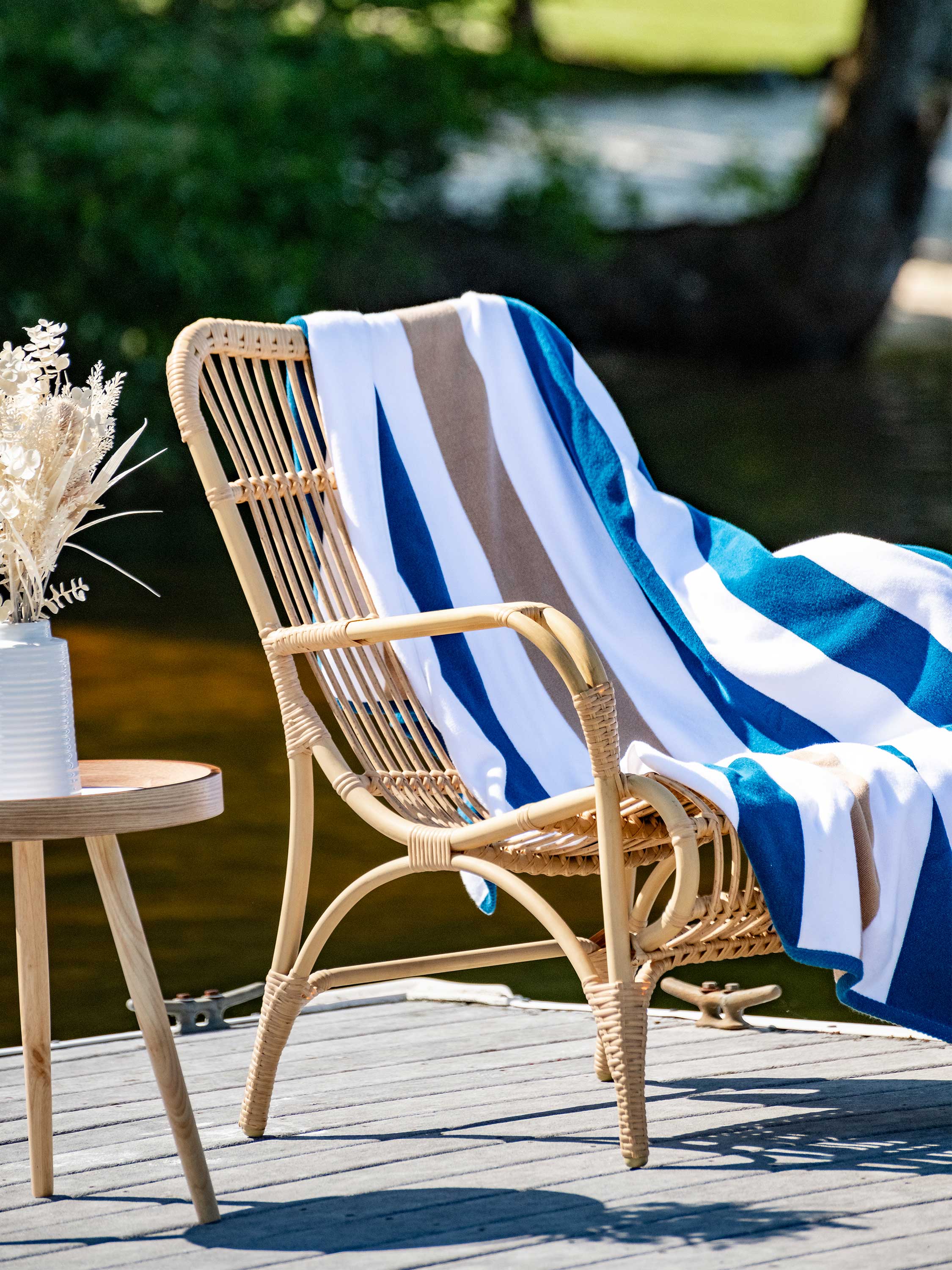 Wicker chair with a striped towel draped over it on a wooden deck by a body of water.