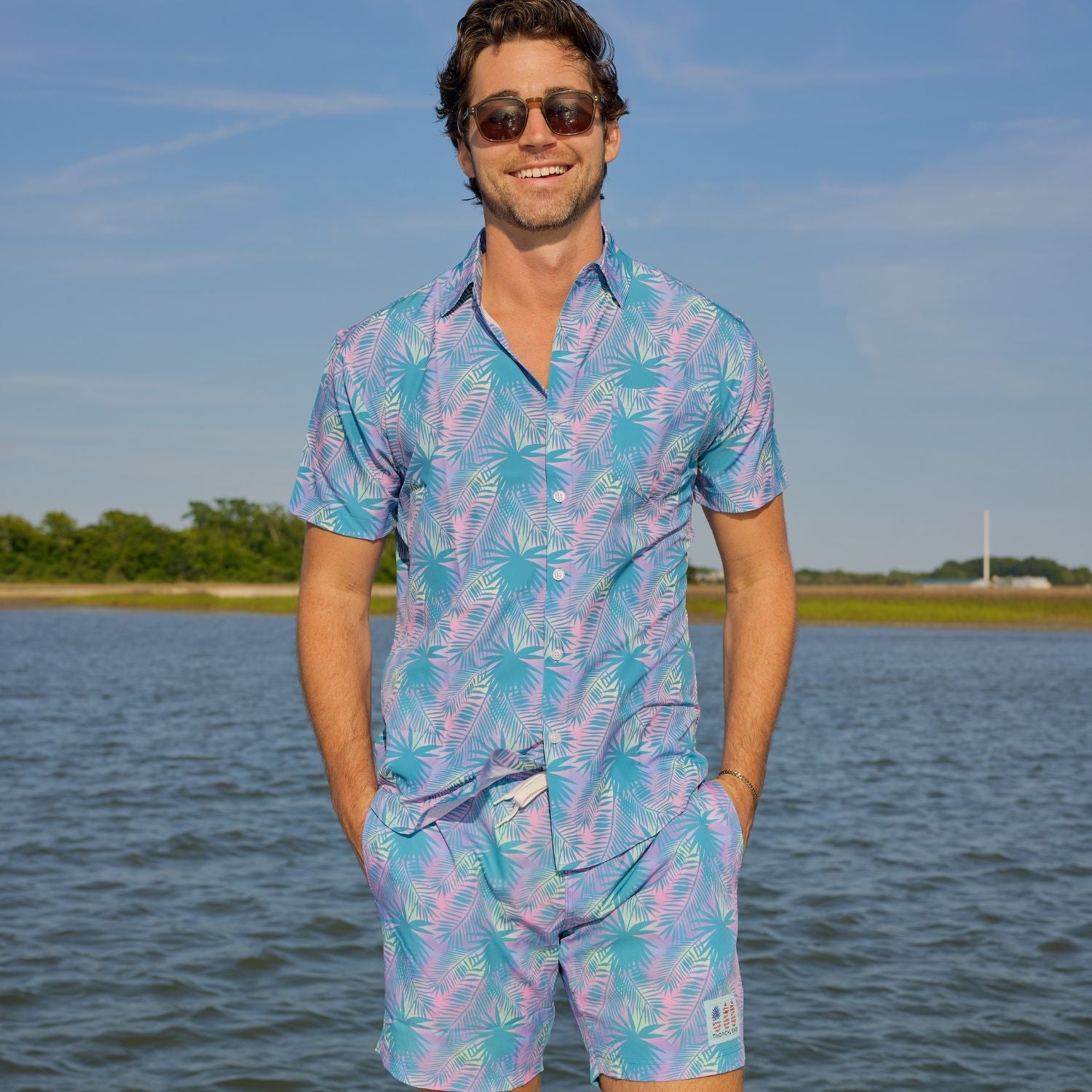 Man wearing a colorful Hawaiian shirt and shorts standing by a body of water.