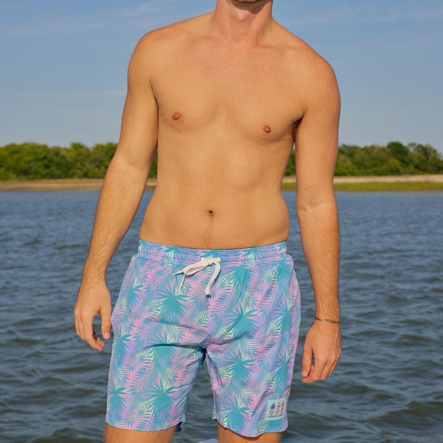 Man wearing colorful swim shorts standing by a body of water with trees in the background