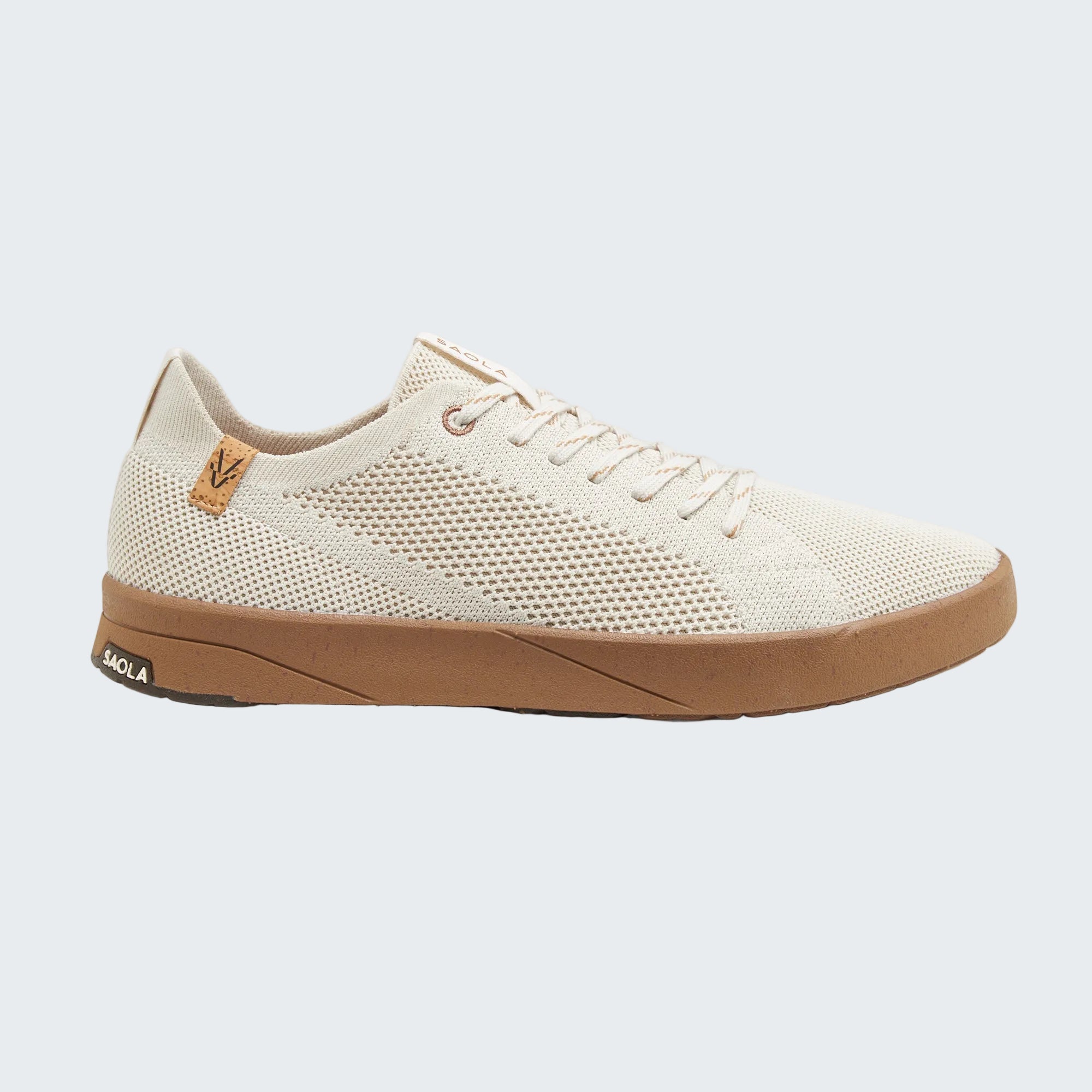A single low-top sneaker shown in a side profile on a light gray background. The shoe features a cream-colored knit upper with a small tan leather logo patch on the heel, a tan midsole, and a black brand name 'SACRA' printed on the heel of the sole.