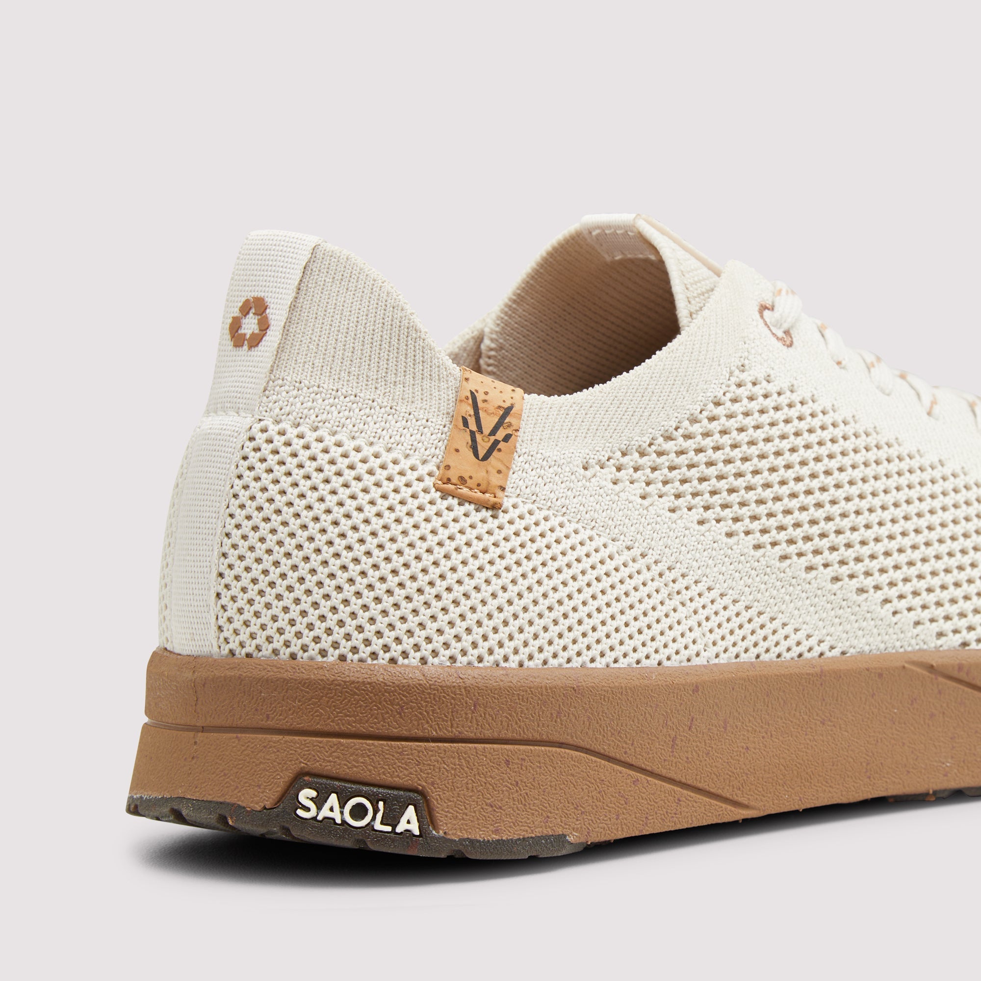 A close-up side view of a knit sneaker in off-white with a tan midsole and a small tan leather logo tab on the side, shown on a light gray background.