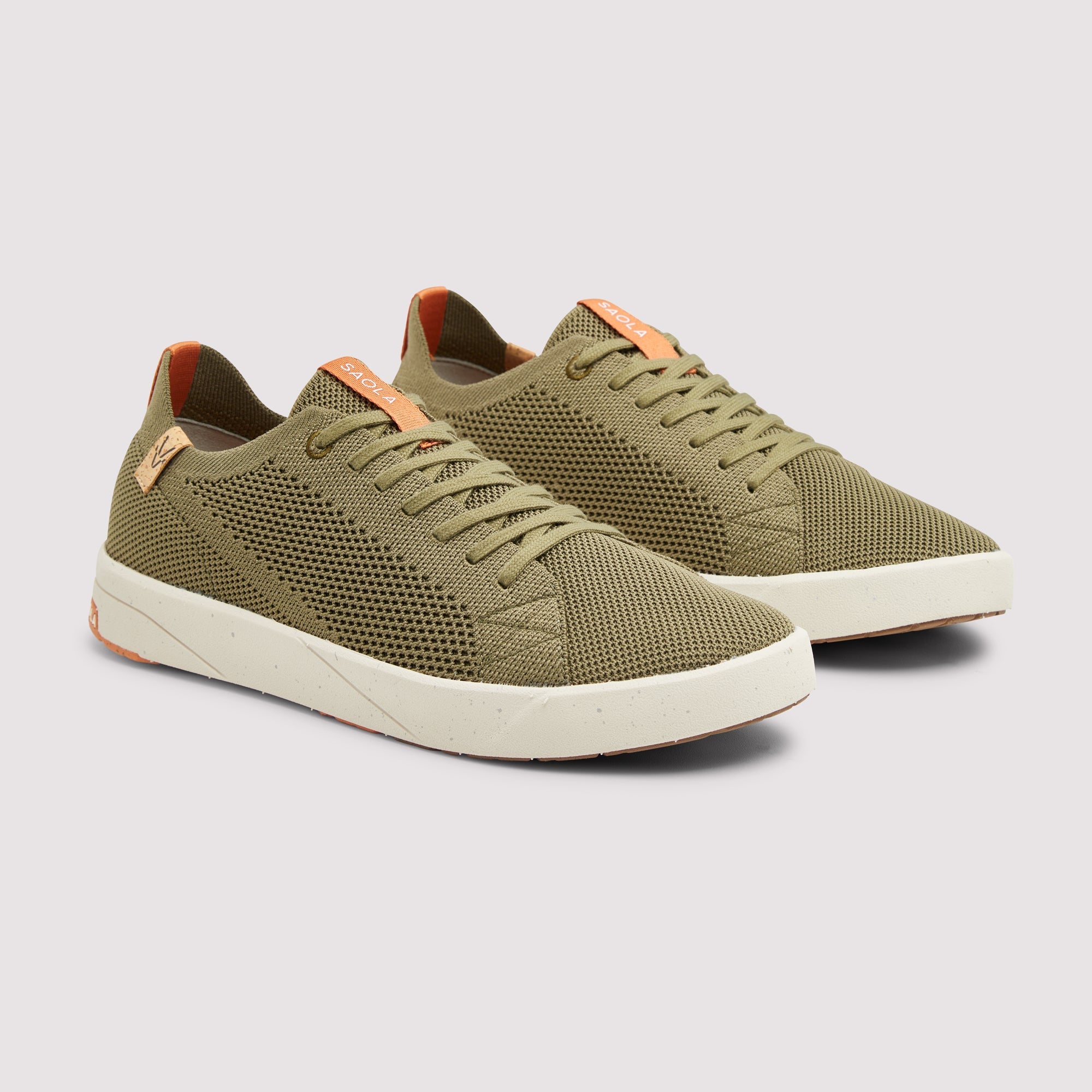 A pair of low-top mesh sneakers in olive green with light orange accents on the tongue and heel tab, and off-white soles, shown on a light gray background.