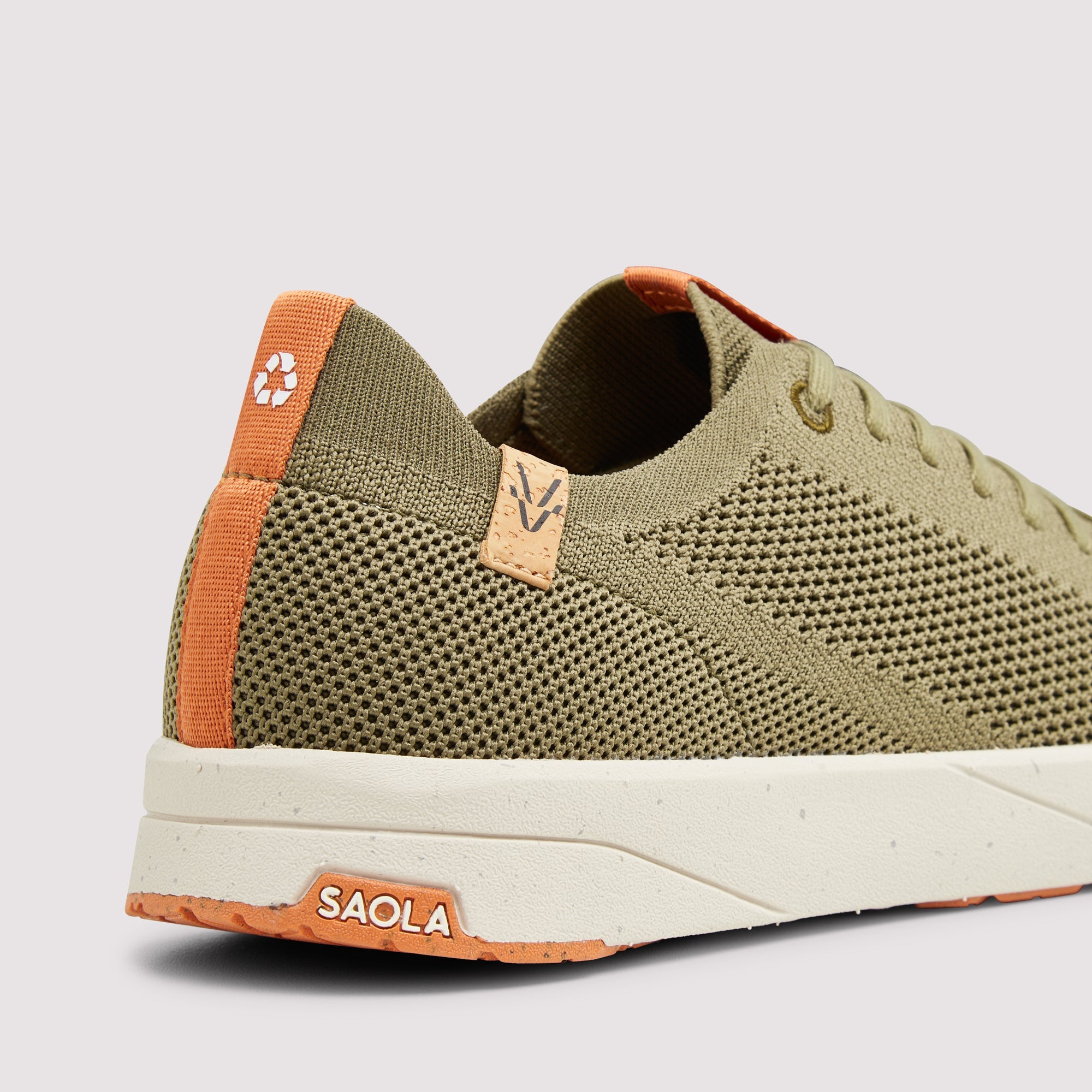 A close-up side view of a knit sneaker in olive green with orange accents on the heel pull tab, tongue detail, and outsole branding, set against a light gray background.