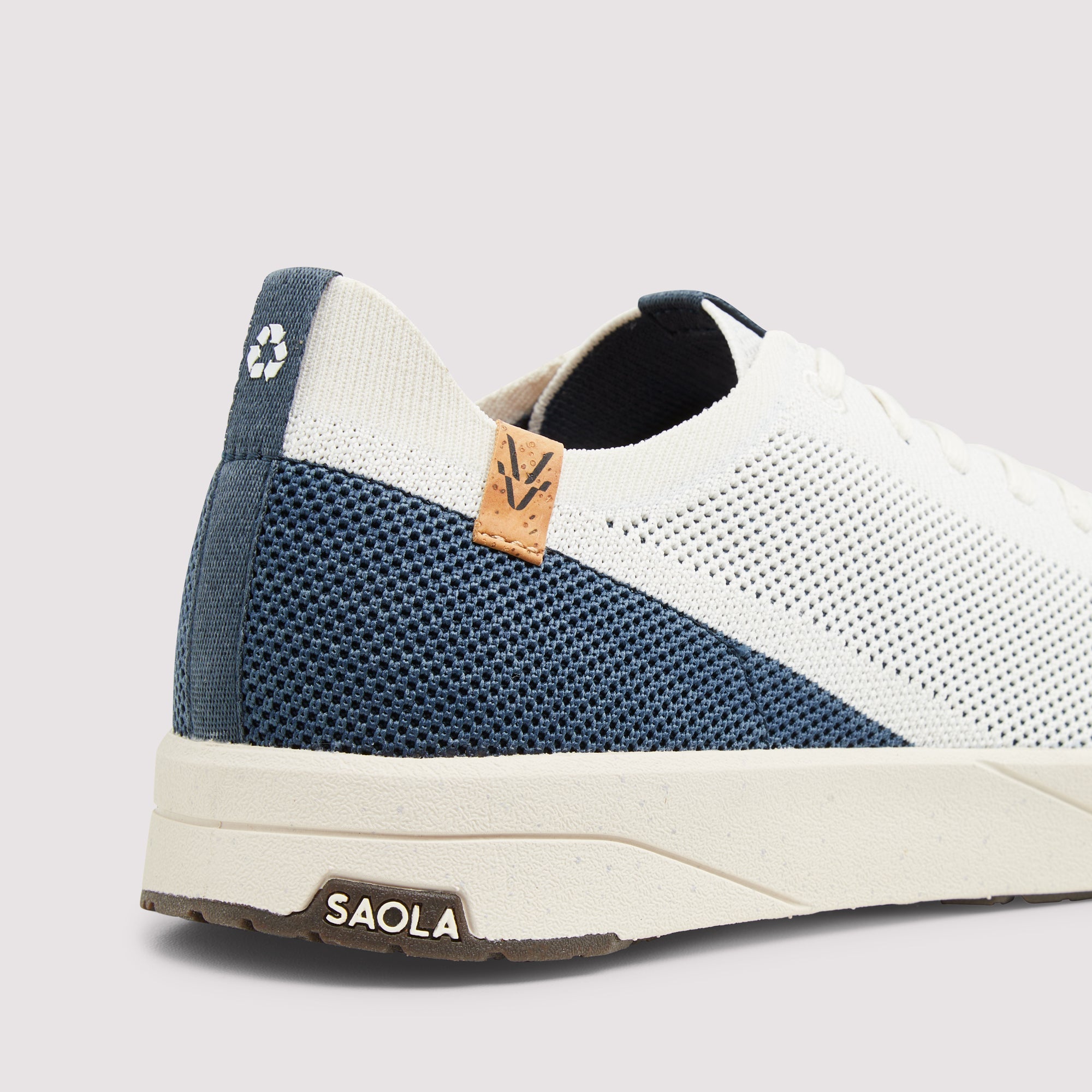 A close-up side view of a low-top sneaker by SAOLA, featuring a white knit upper, a navy blue mesh heel panel, a light brown leather logo tab, and a cream-colored midsole.