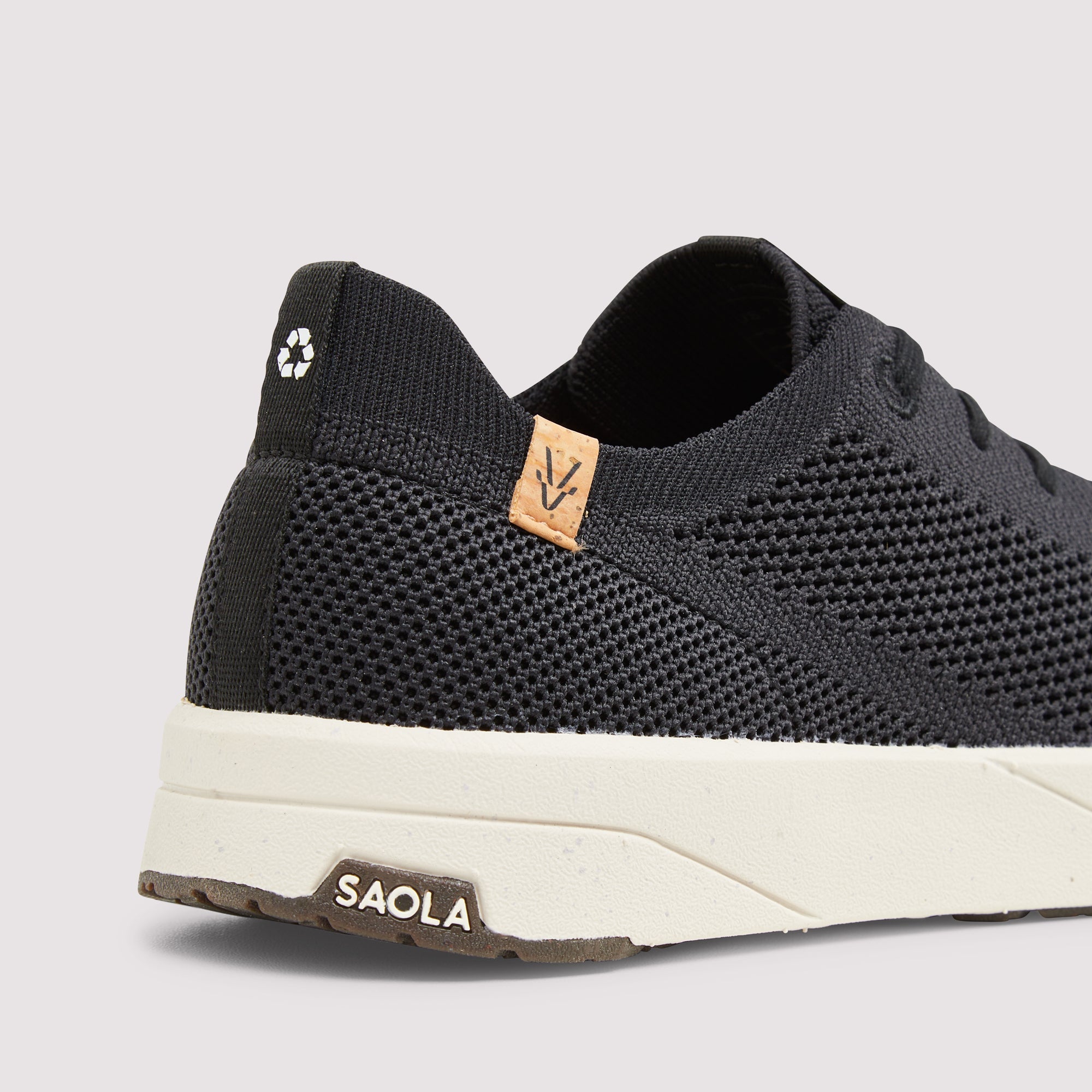 A close-up of a black knit athletic sneaker with a thick off-white midsole and a tan leather heel tab featuring a stylized 'V' logo. The brand name 'SAOLA' is printed on the outsole.