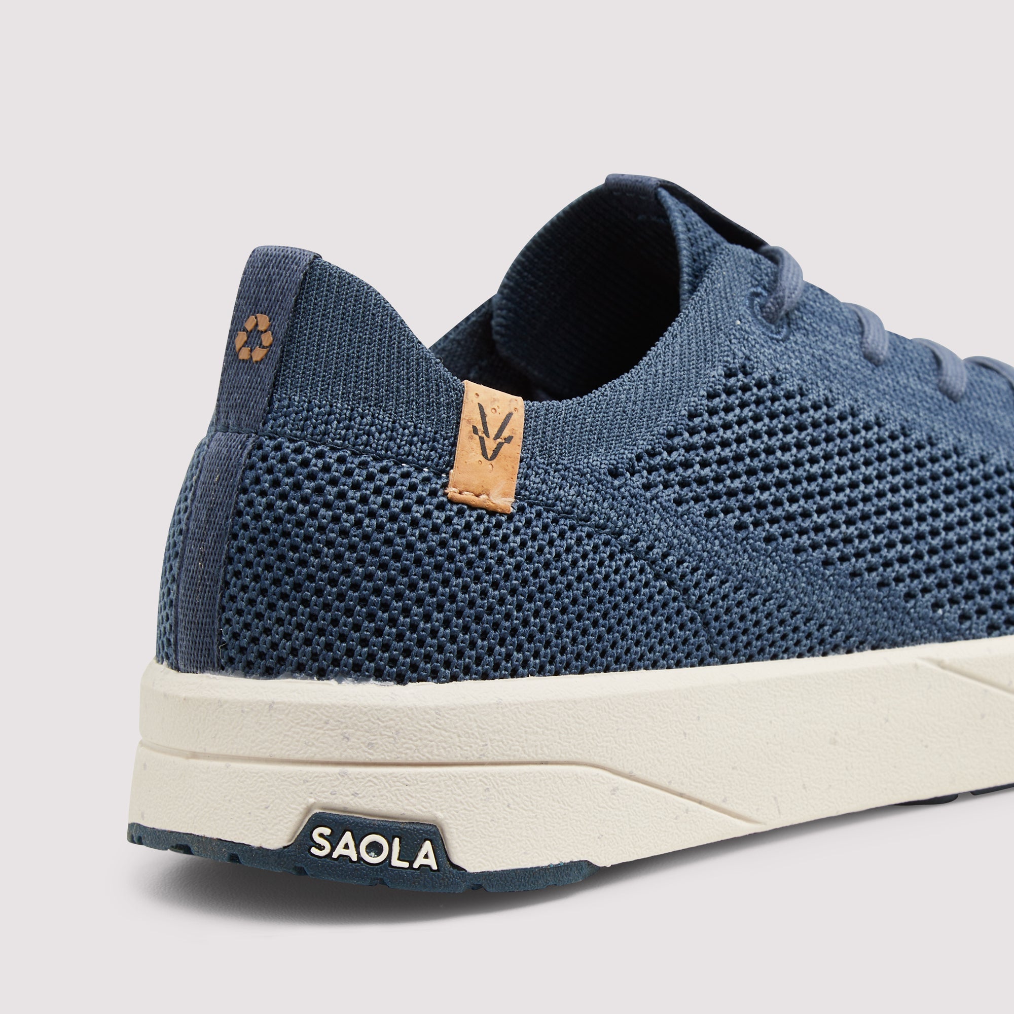 A close-up shot of a blue knit sneaker by Saola, showing the textured knit upper, a small tan leather logo tab, and a thick off-white midsole on a light gray background.