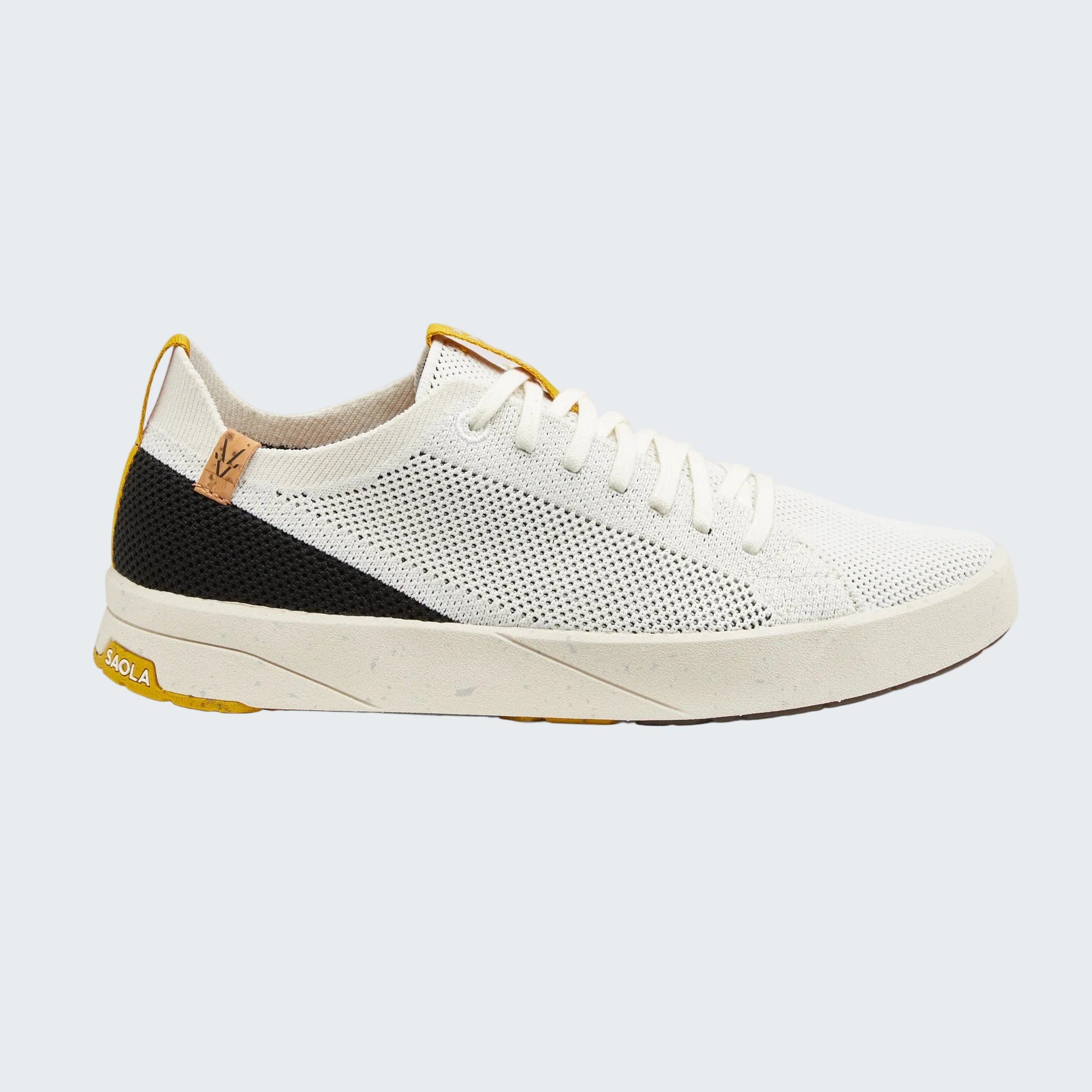 A white knit sneaker with a black side panel, white laces, and yellow accents on the heel tab, tongue, and outsole, shown against a light gray background.