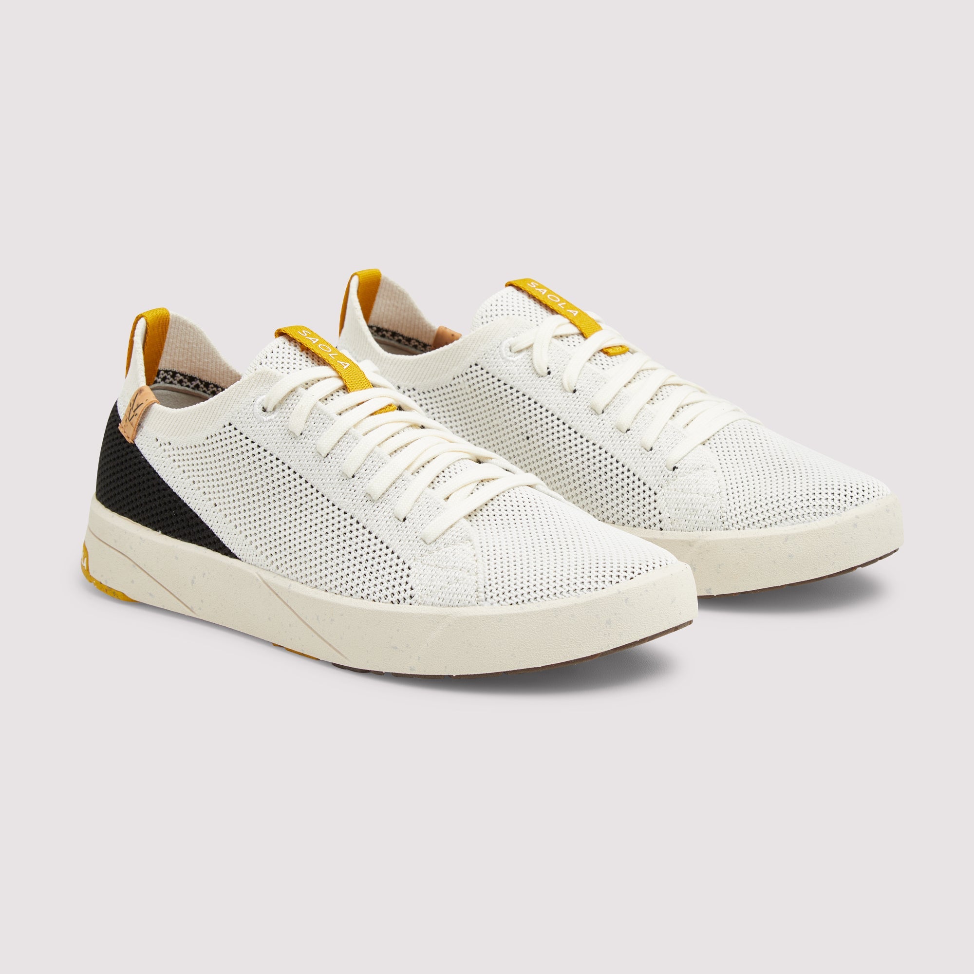 A pair of white low-top sneakers with a perforated mesh upper, white laces, a black heel accent, and yellow pull tabs, shown on a light gray background.