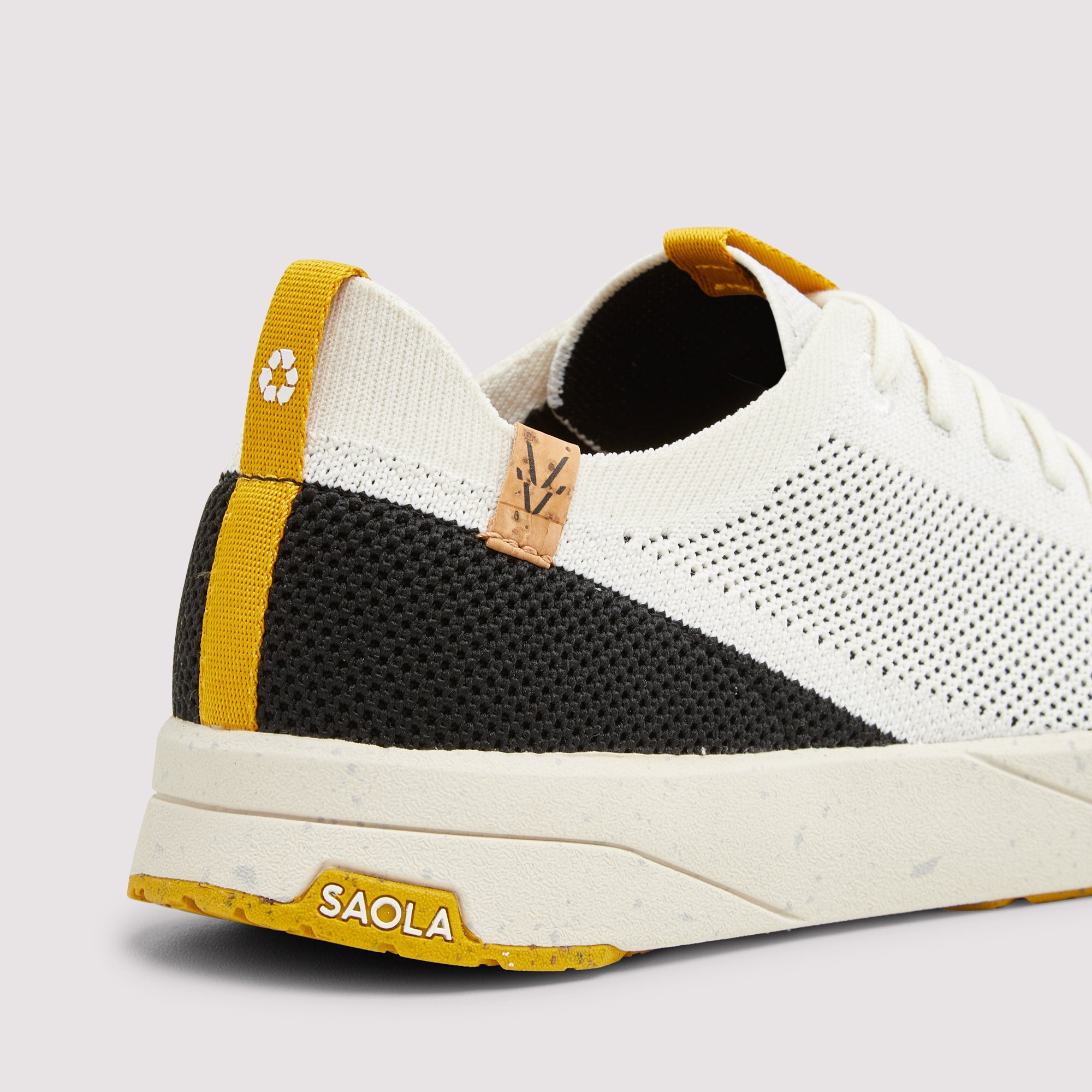 A close-up rear three-quarter view of a white, black, and yellow knit sneaker. The shoe features a white knit upper, a black knit panel, a yellow heel pull tab with a recycling symbol, and a yellow outsole.