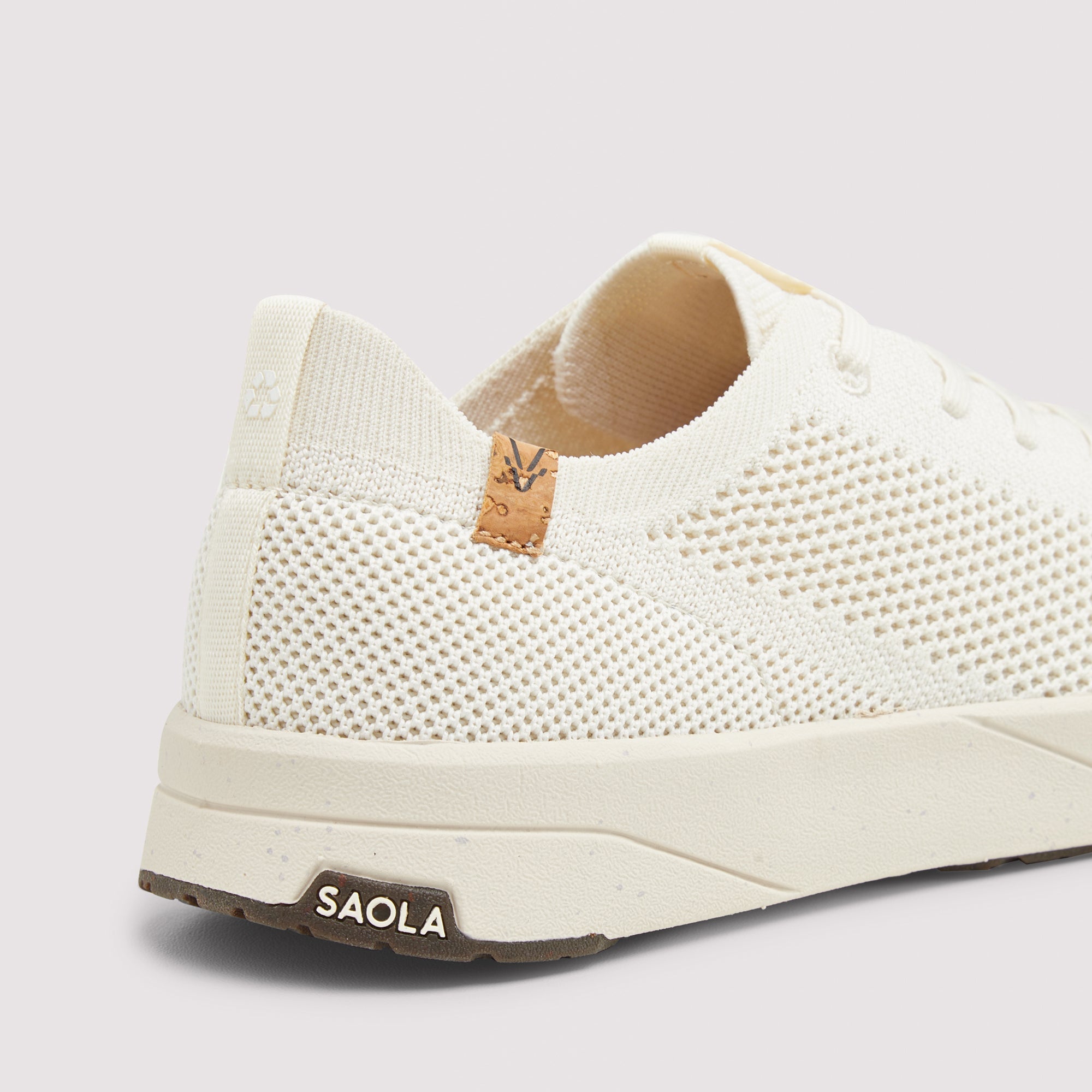 Close-up of the heel and side of a knit slip-on sneaker in off-white/cream with a tan pull-tab detail and a dark brown sole edge. The brand name SAOLA is visible on the heel tab.