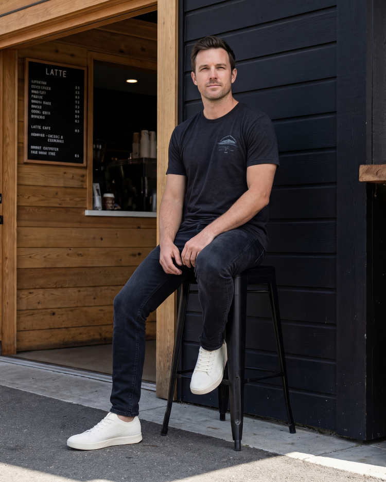 Man sitting on a stool outside a cafe wearing a black t-shirt and jeans.