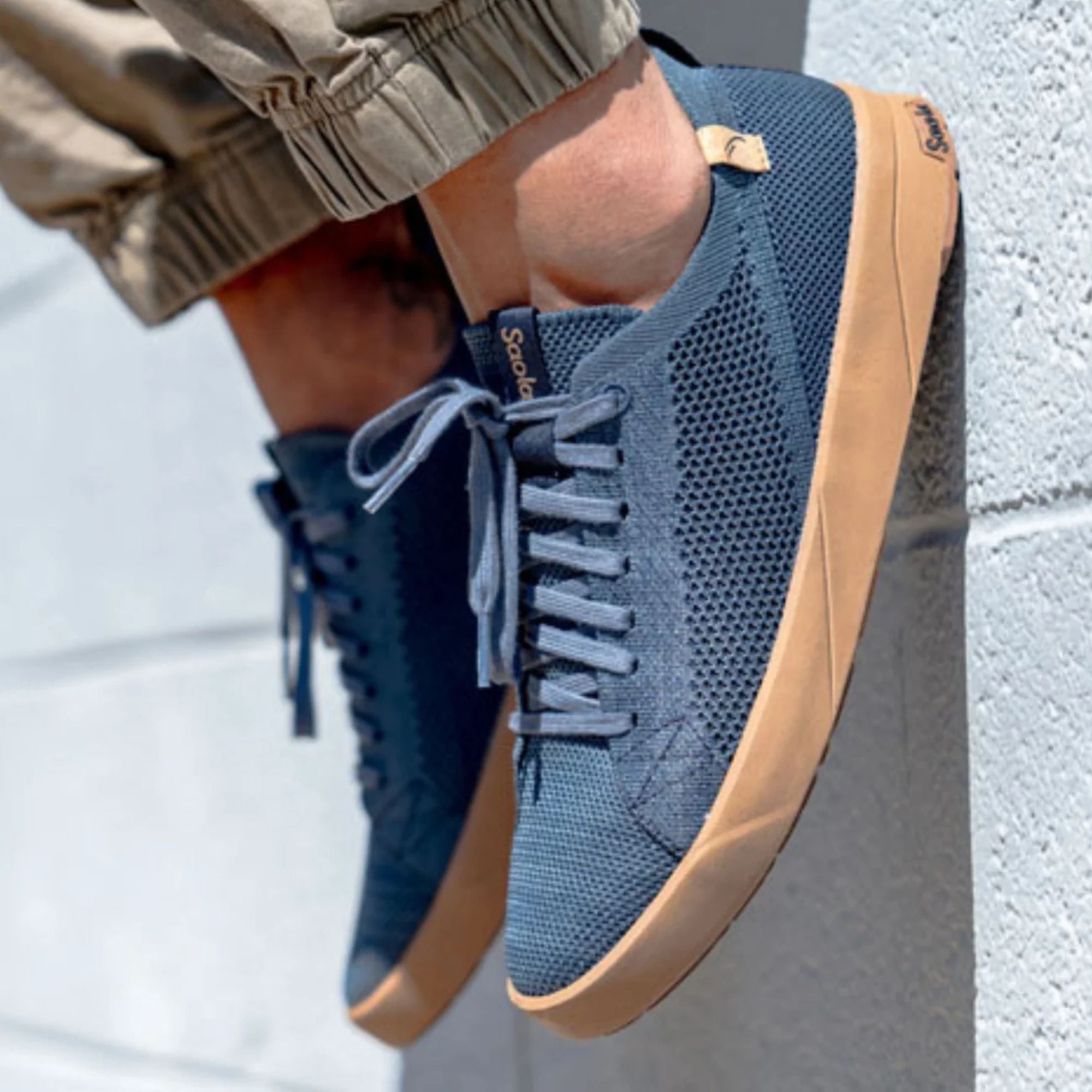 A close-up of a person wearing blue mesh sneakers with tan soles, photographed against a light gray textured wall. The brand name 'sodapop' is visible on the tongue tag.