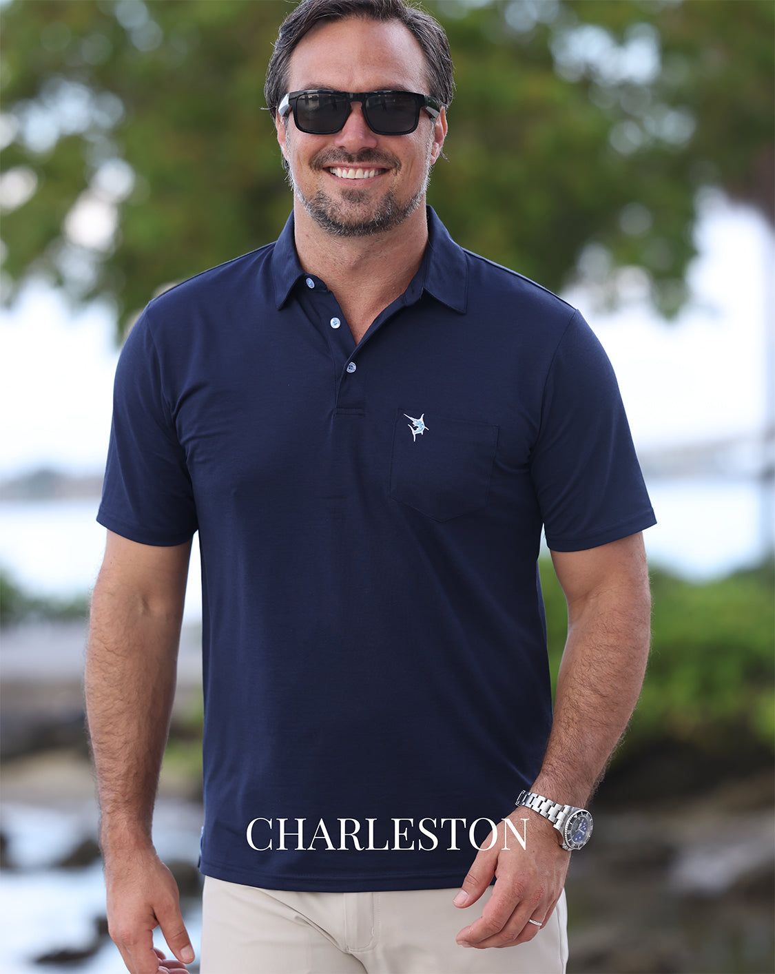 Man wearing a navy polo shirt with a logo, sunglasses, and a watch, standing outdoors.